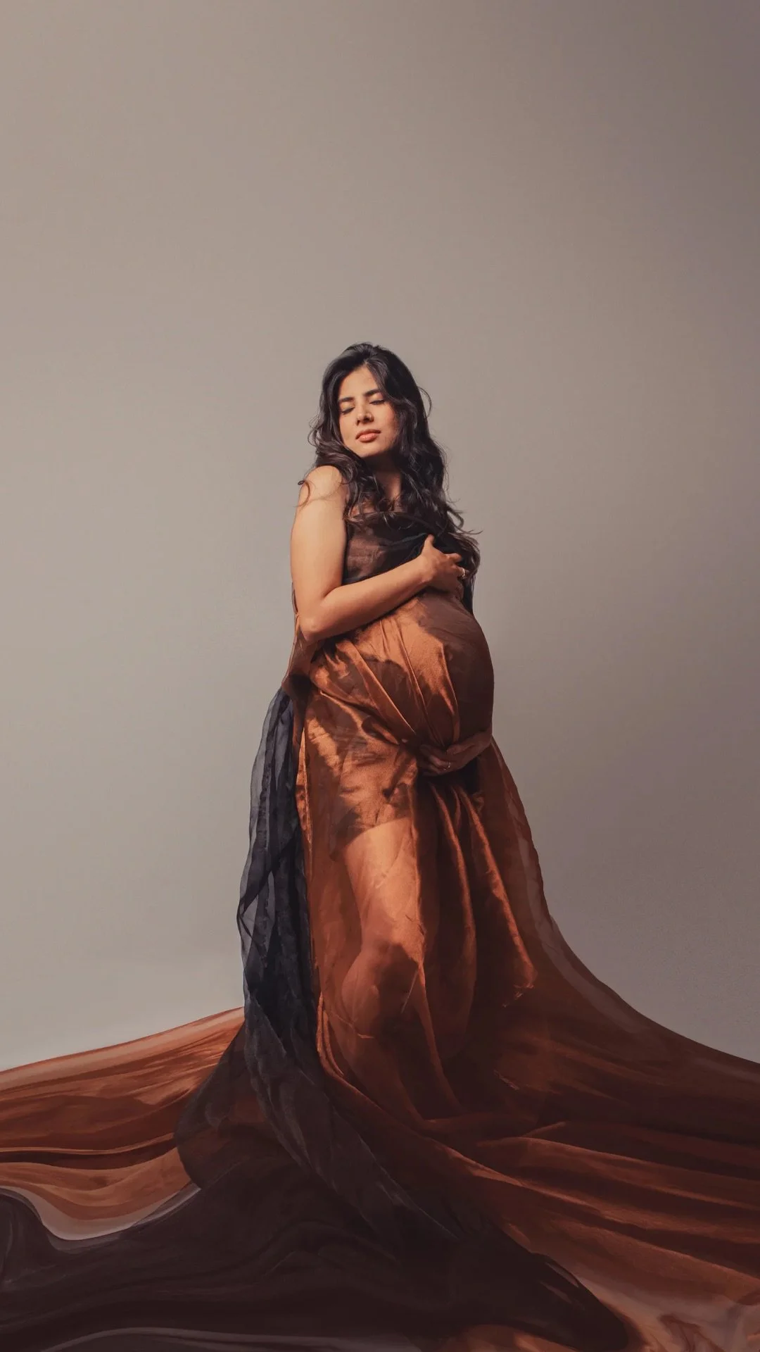 A pregnant woman with long dark hair is wearing a flowing, sheer dress with shades of brown and black. She stands against a plain background, holding her belly, with a serene expression.