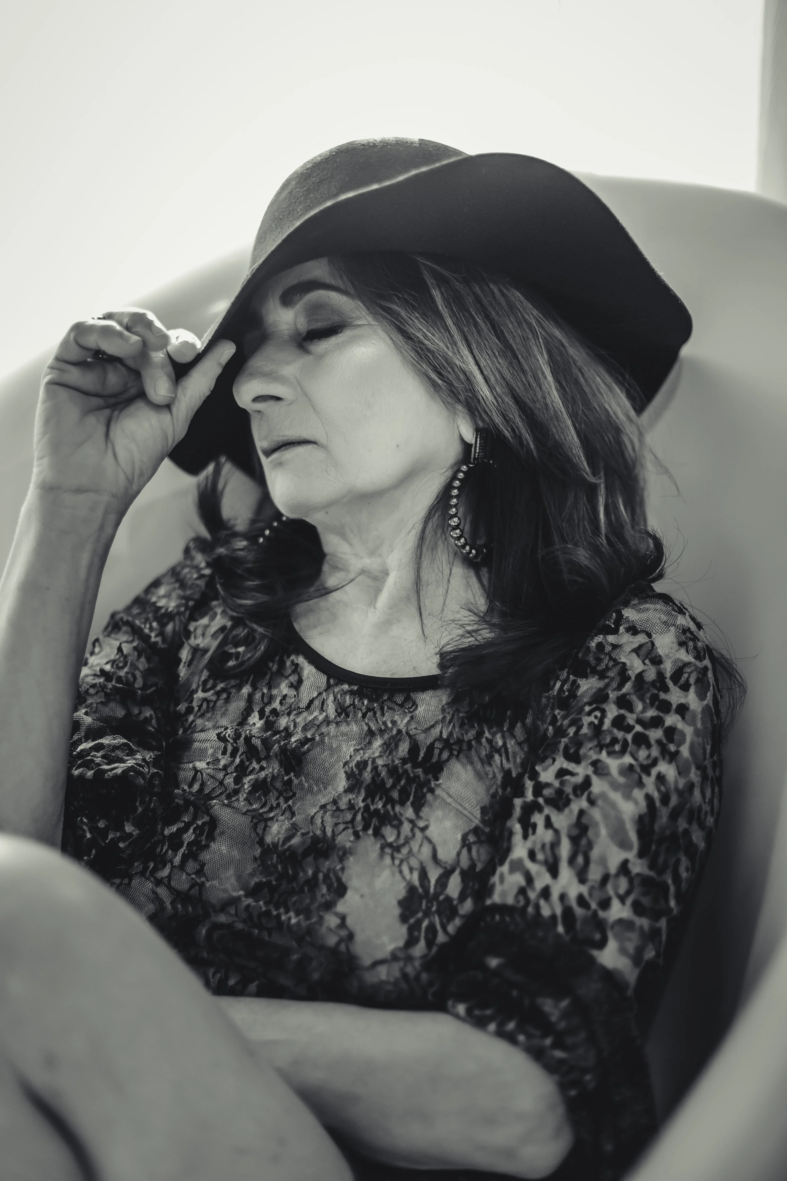 Black and white photo of a woman with long hair wearing a large floppy hat, lace patterned top, and hoop earrings, resting with her eyes closed and her hand holding the brim of her hat.