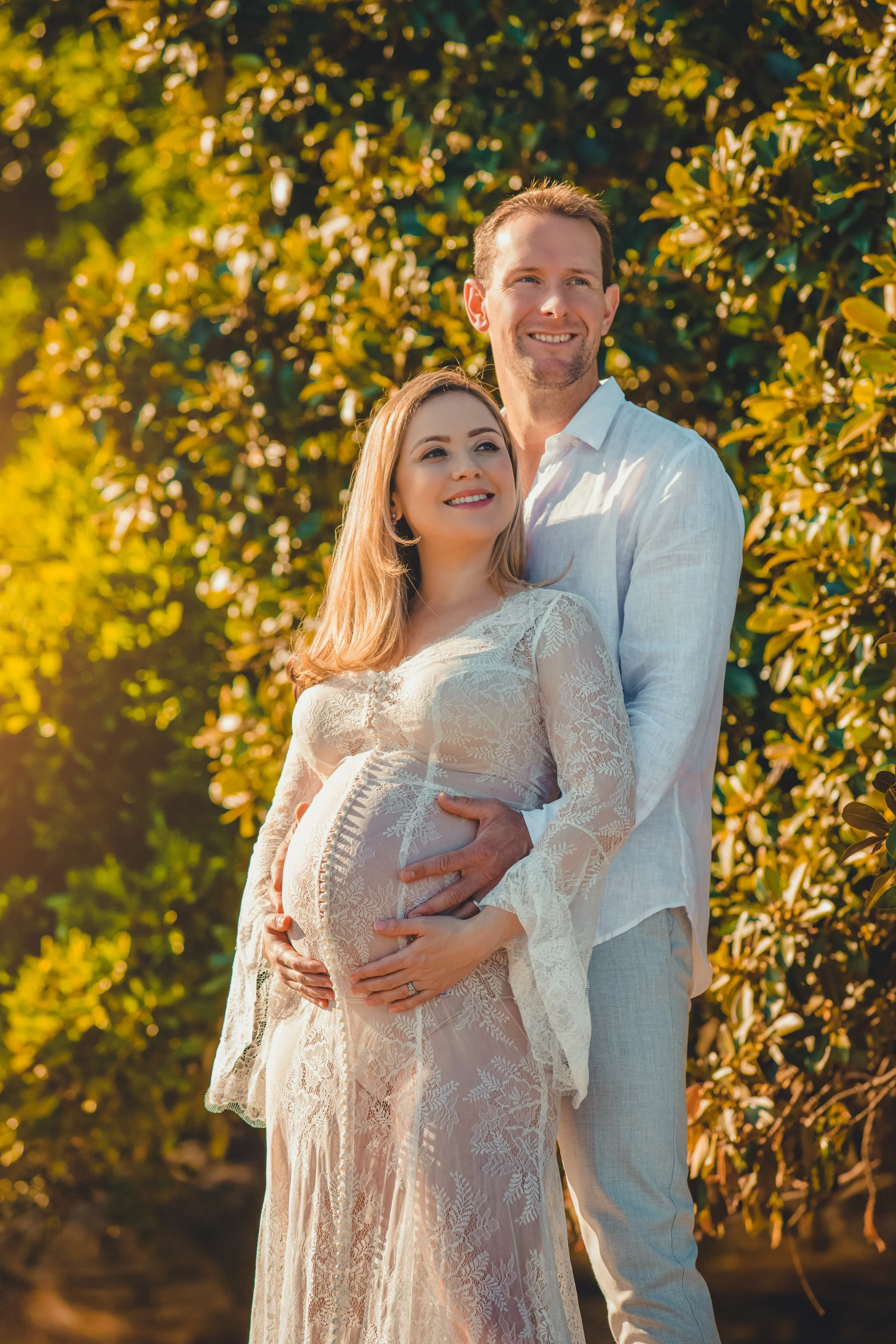 A pregnant woman in a sheer lace dress and a man in a white shirt pose outdoors among greenery with warm sunlight. The man holds the woman from behind, both smiling and looking into the distance.