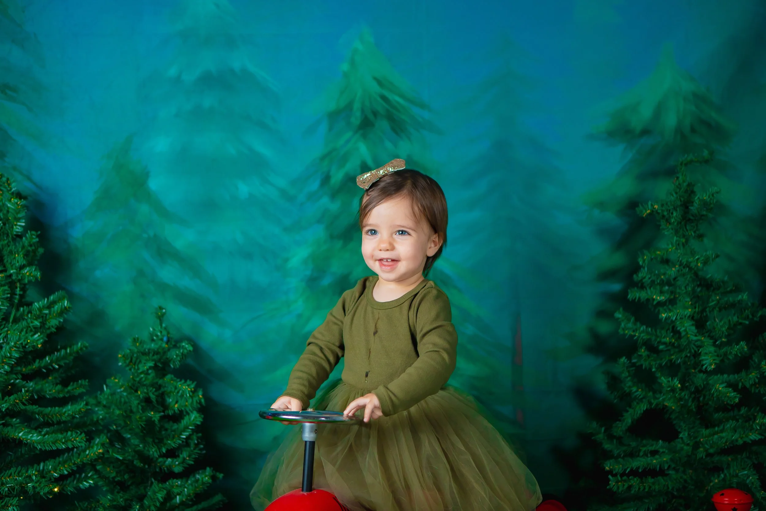 A young girl with brown hair, wearing a green top and a green tulle skirt, is sitting on a red ride-on toy in front of a painted forest backdrop with pine trees. She has a glittery gold bow in her hair and is smiling.
