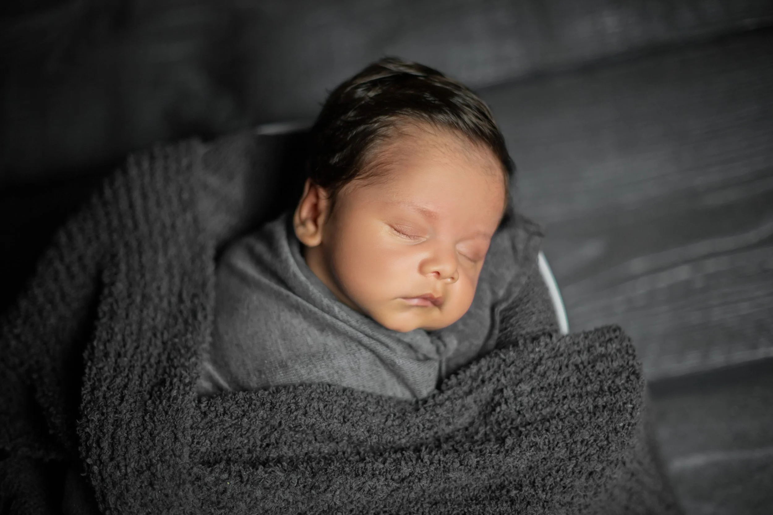 A close-up of a sleeping newborn baby wrapped in a dark grey blanket.