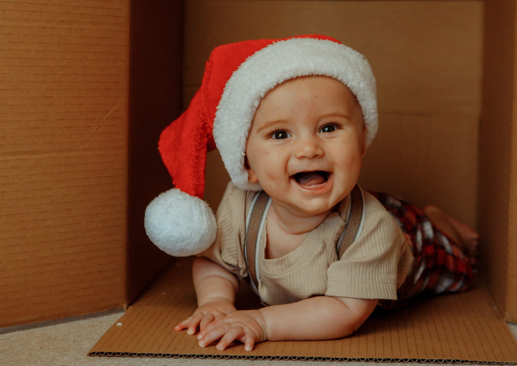 A baby wearing a Santa hat, smiling, crawling on a cardboard surface in a small enclosed space.