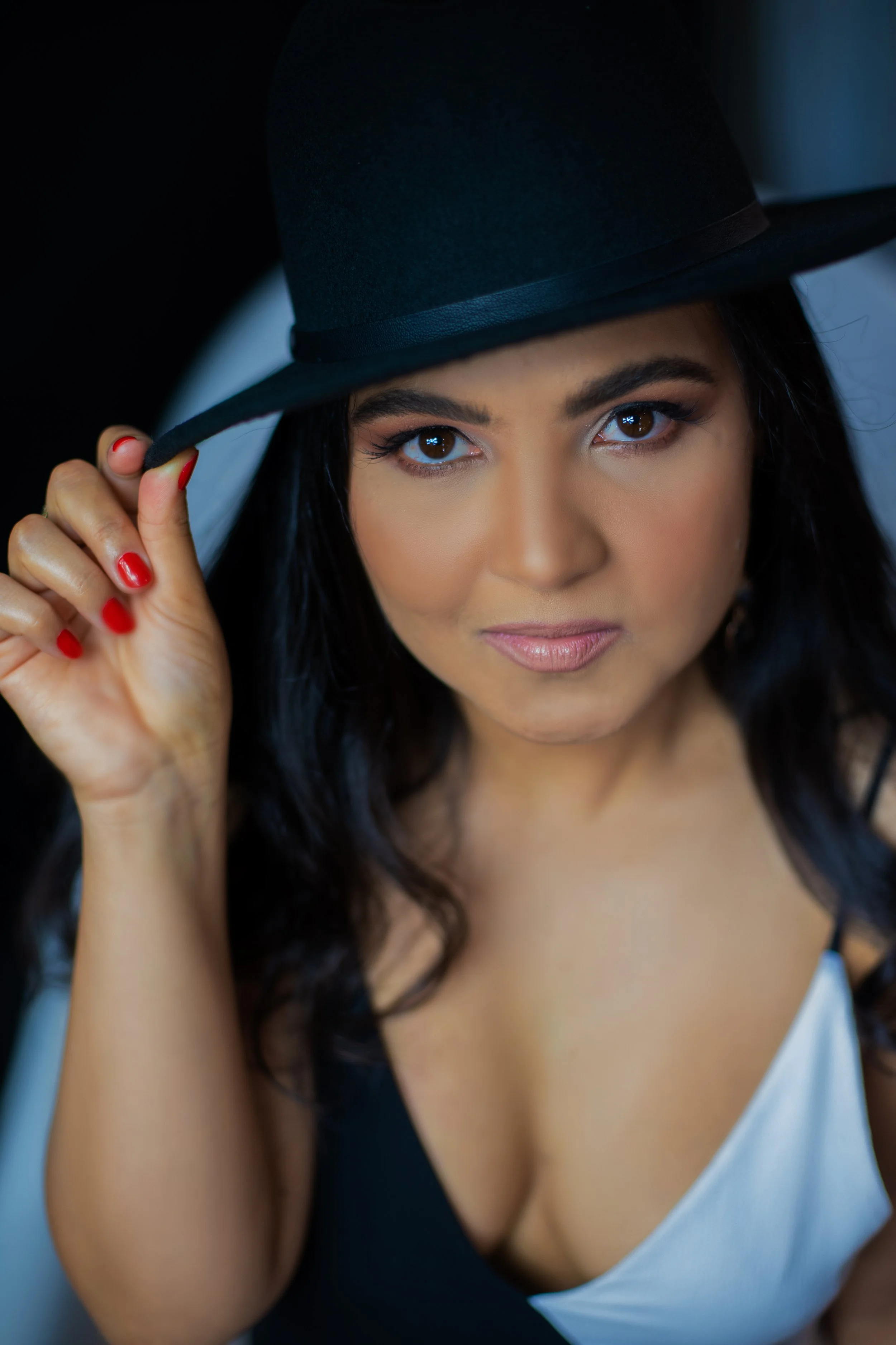 A woman with dark hair and brown eyes wearing a black hat, holding the brim with her right hand, and looking into the camera.