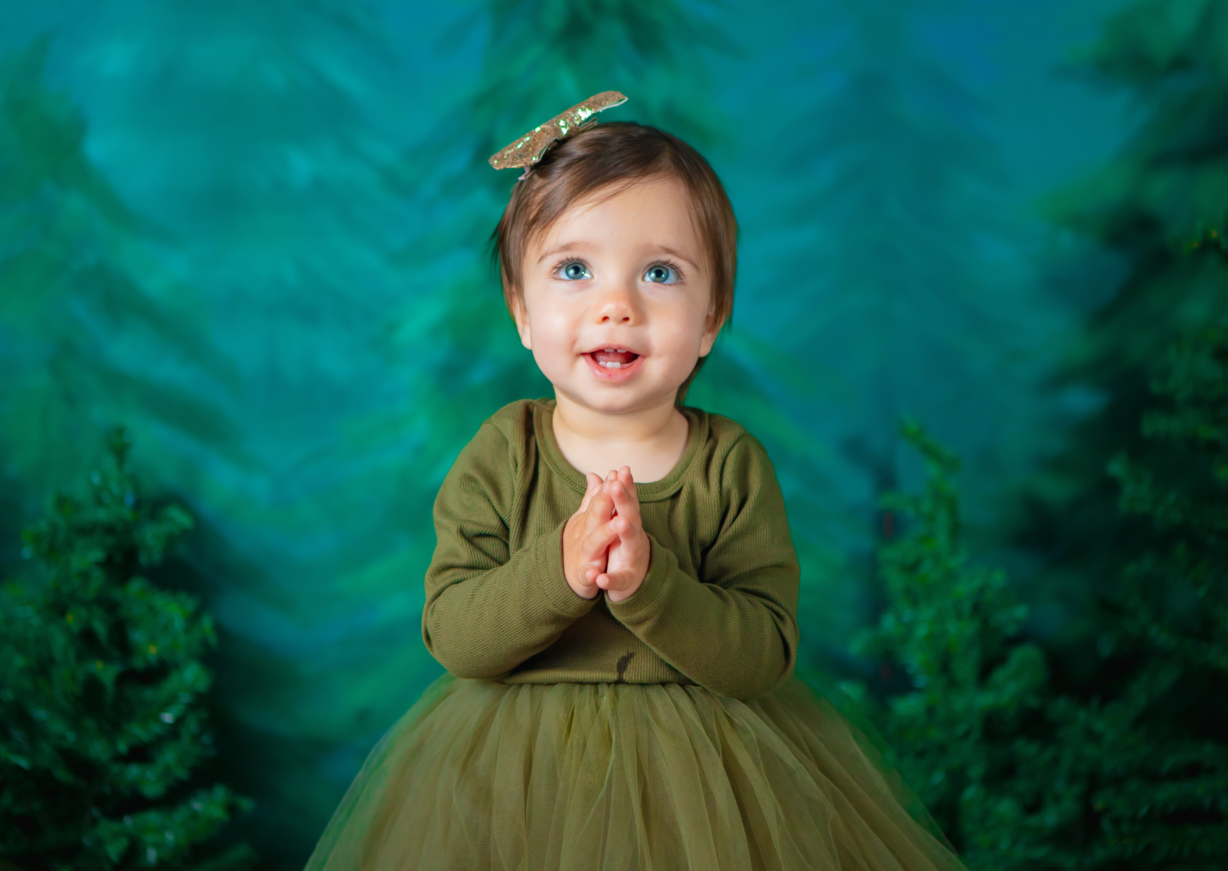 A young girl with blue eyes and brown hair wearing a green long-sleeve shirt and tulle skirt, with her hands clasped together, in front of a green and blue brushed background, looking up with a smile.
