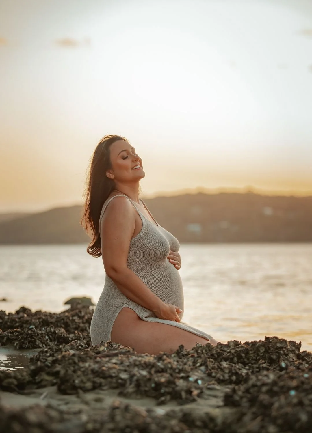 Pregnant woman kneeling on rocky shoreline at sunset, smiling with eyes closed, wearing a beige tank top, with water and hills in the background.