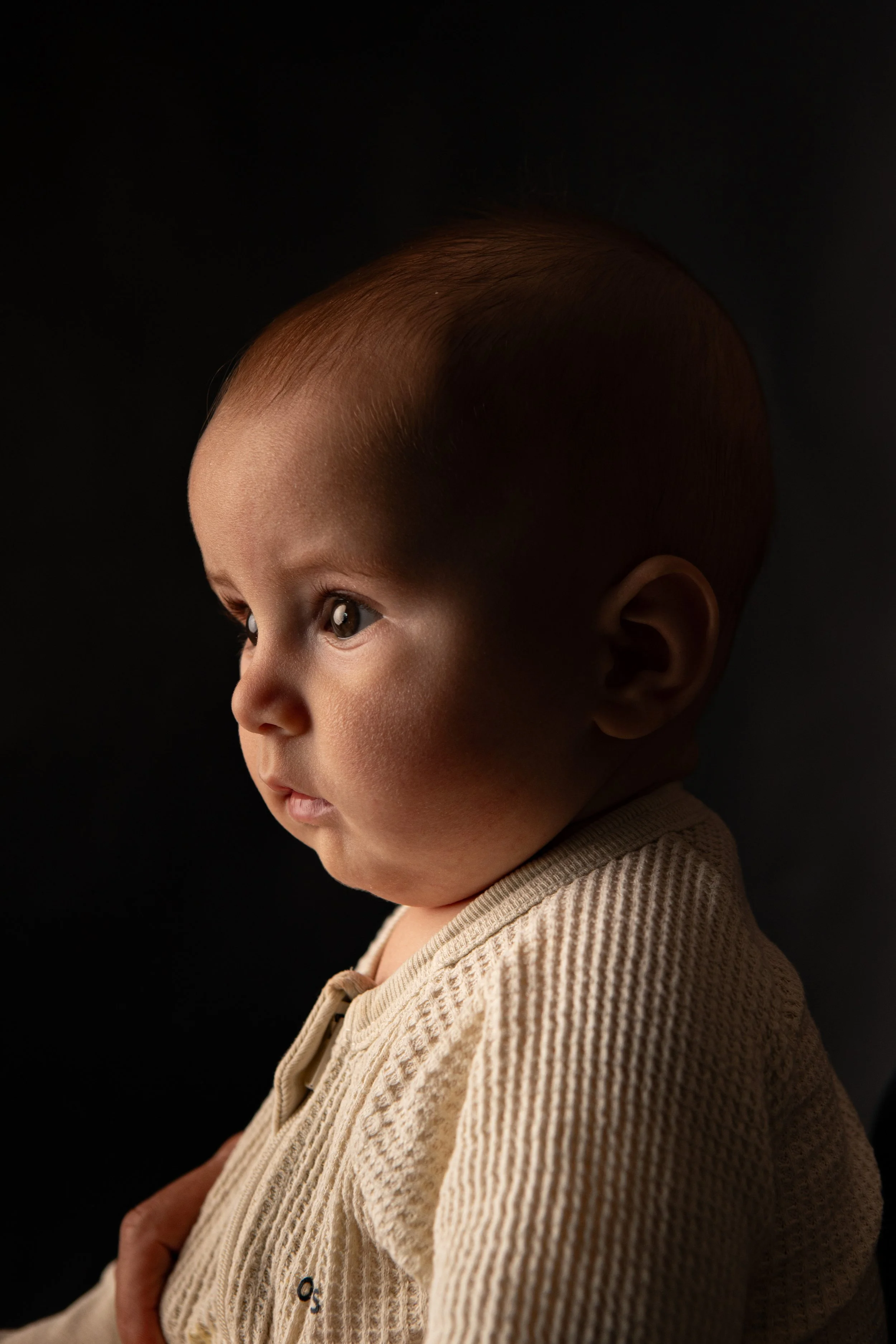 Side profile of a young child with short hair and fair skin, wearing a beige textured shirt, against a dark background.