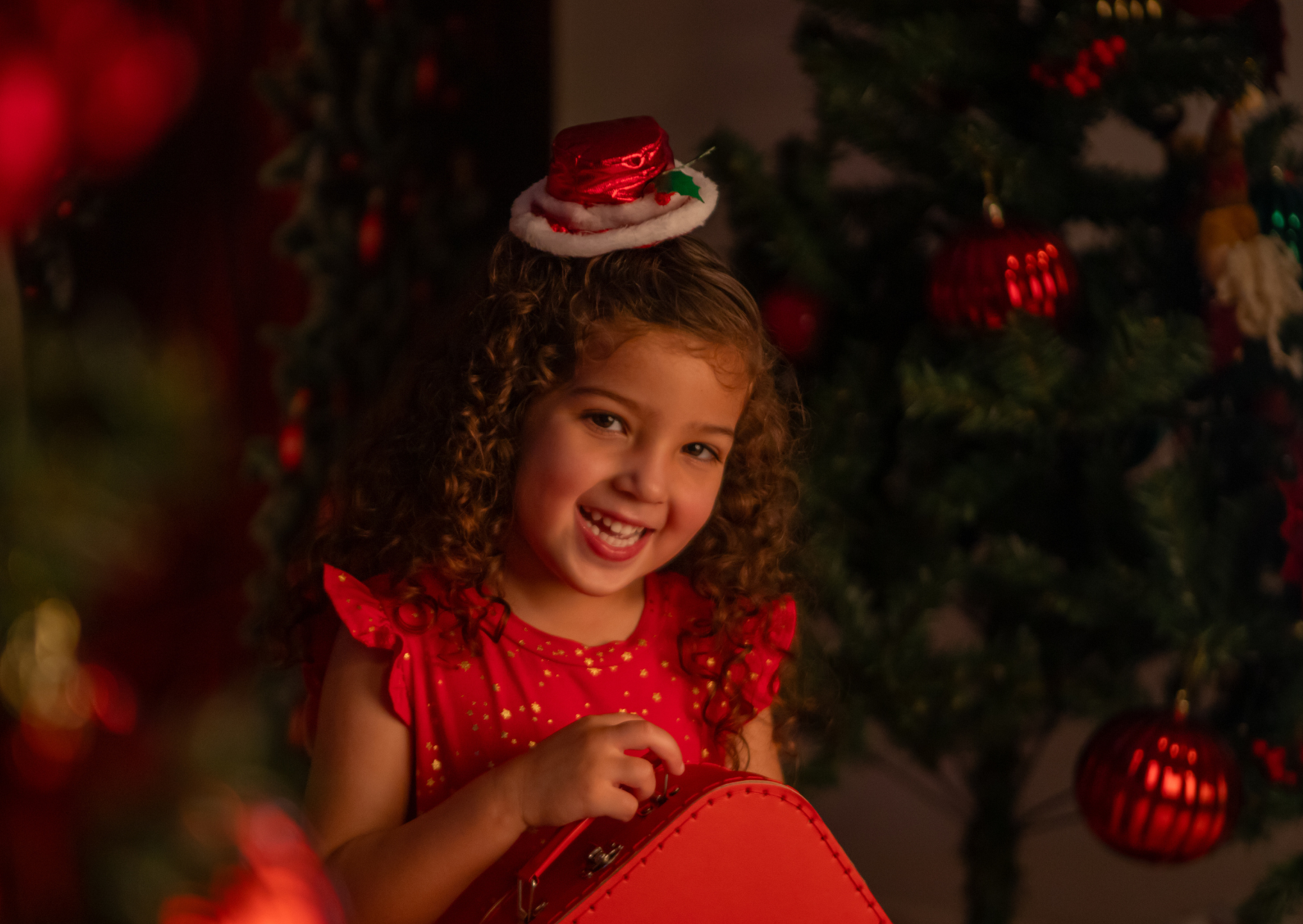 A young girl with curly hair, wearing a red dress with gold accents, is smiling while holding a red suitcase in front of a decorated Christmas tree. She has a small Santa hat on her head and is surrounded by festive ornaments.