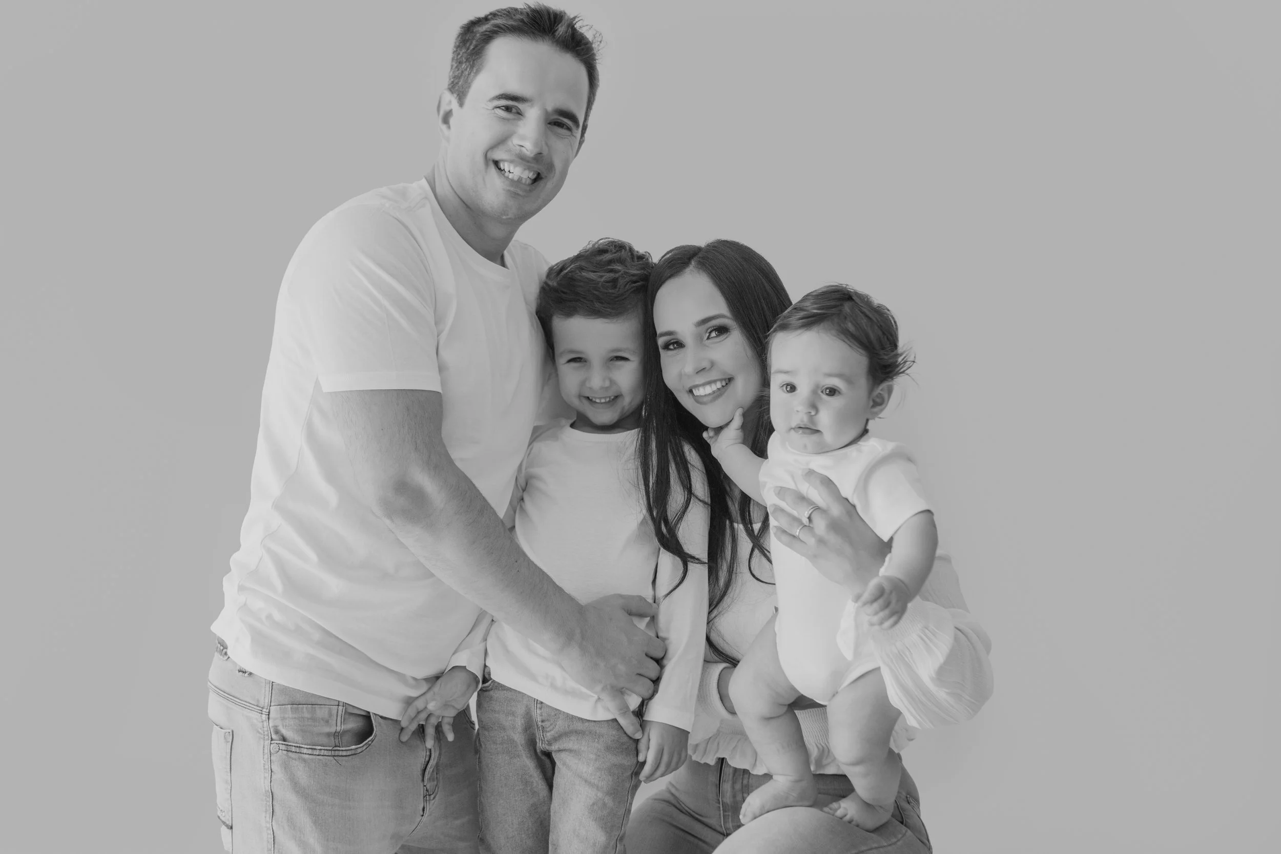 Black and white photo of a smiling family of four with a man, woman, and two young children, posing together against a plain background.