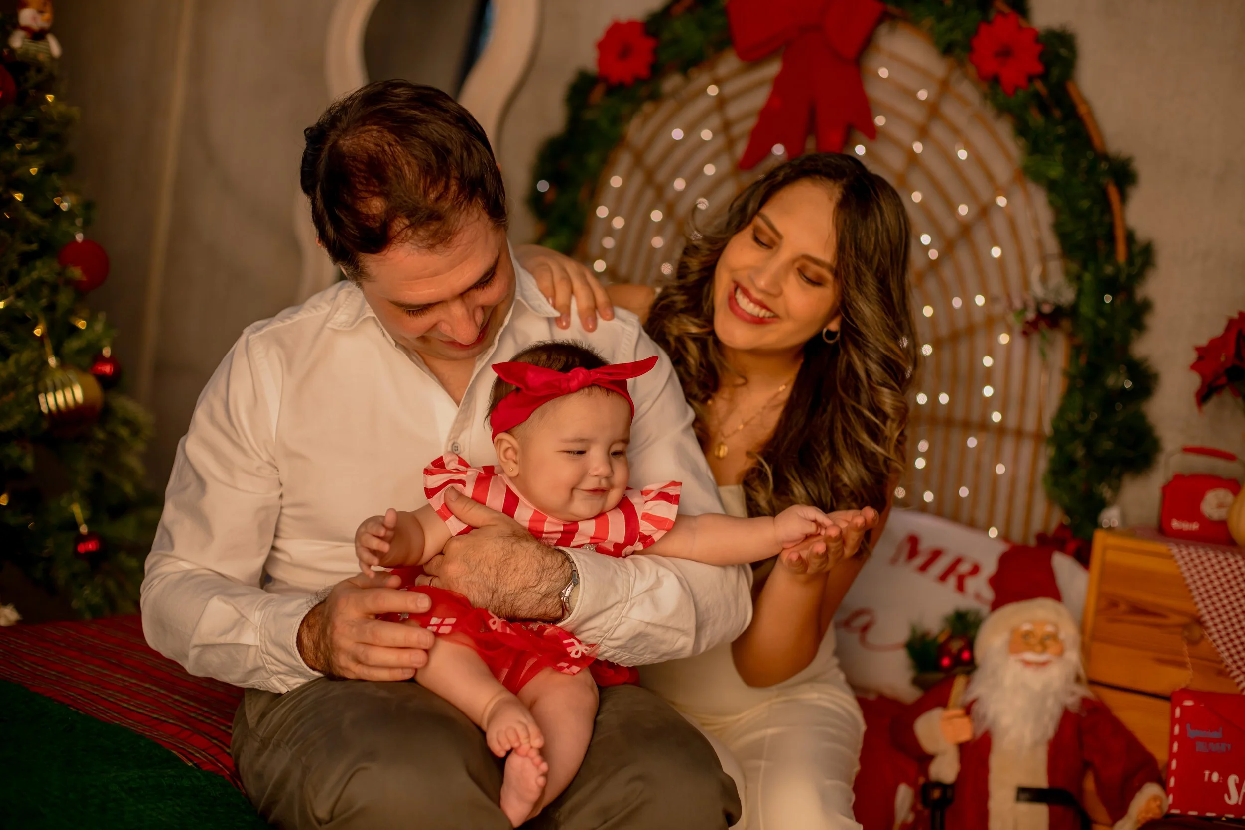 Family celebrating Christmas indoors, with a father, mother, and baby girl smiling and playing together near decorated Christmas tree with lights and ornaments, in front of a festive backdrop with holiday decorations.