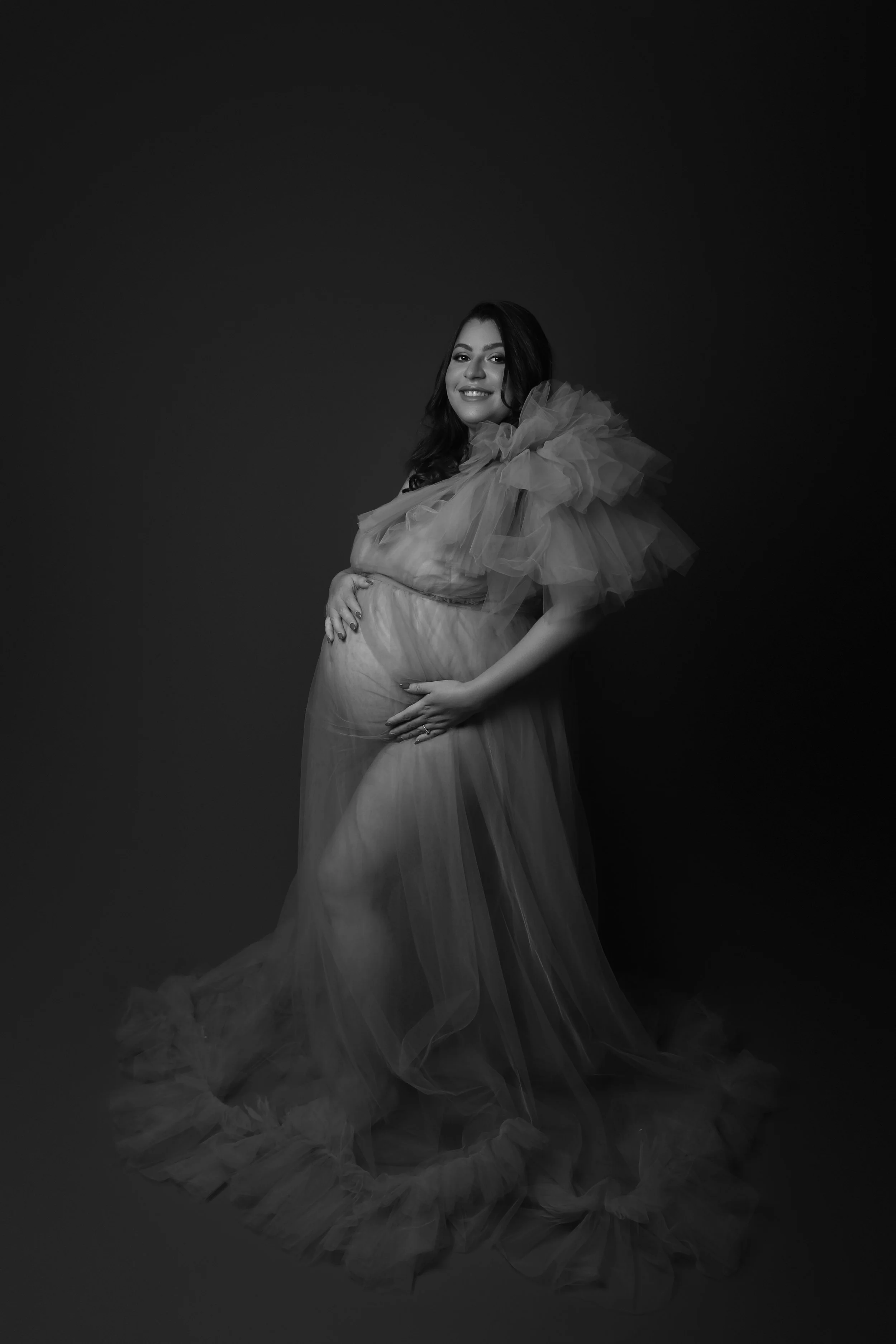 Black and white portrait of a pregnant woman wearing a flowing gown with ruffled sleeves, standing against a dark background and smiling while gently holding her belly.