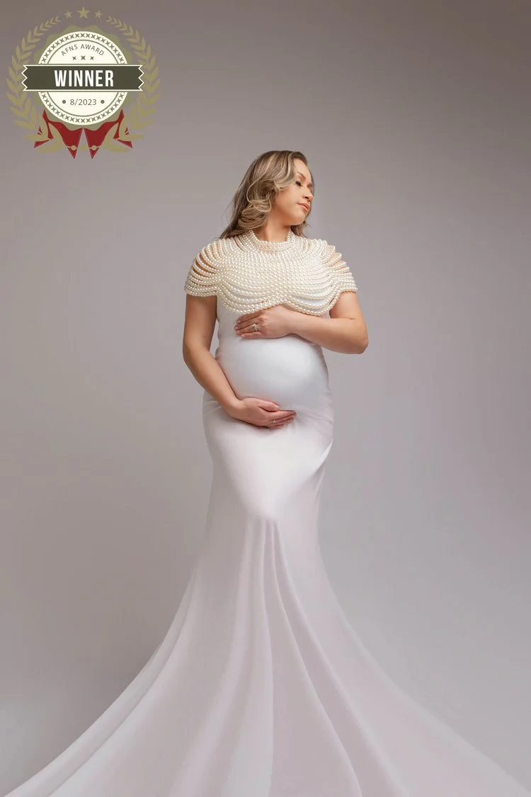Pregnant woman wearing a white gown with a pearl-embellished cape, standing against a plain background. There is a gold and red ribbon badge in the top left corner indicating she is a winner of an award in August 2023.