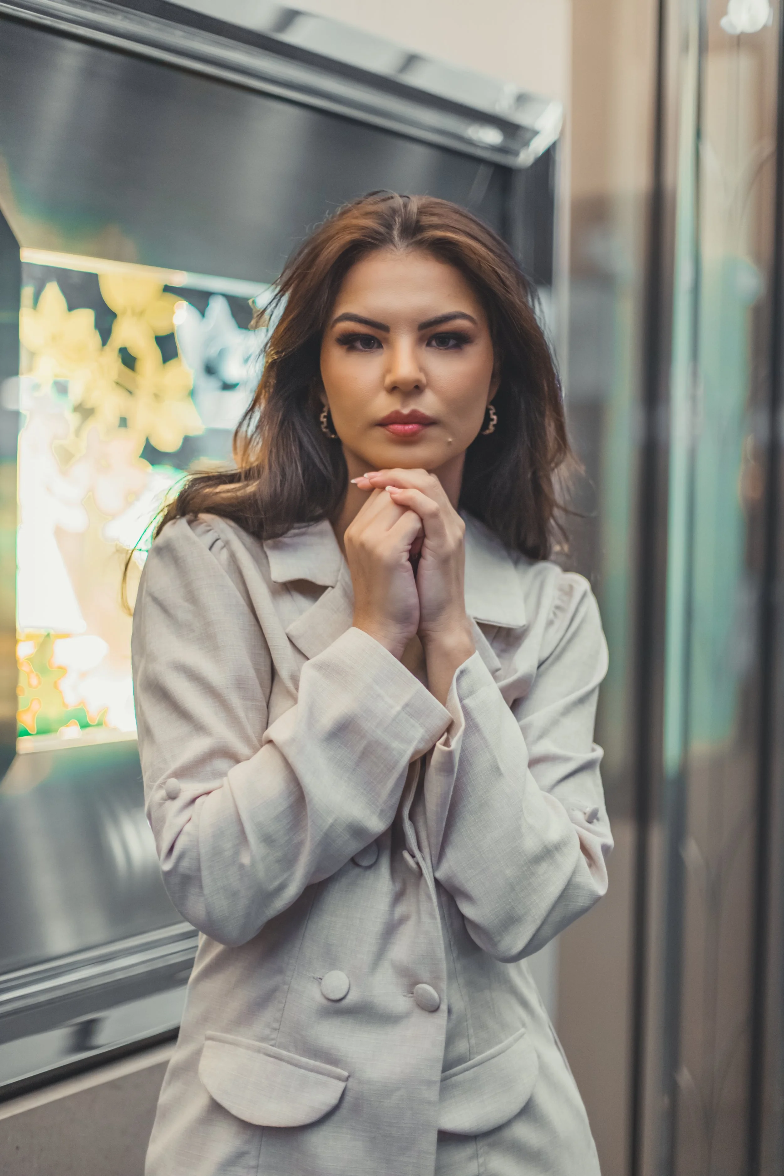 A woman with brown hair, wearing a beige jacket, stands with her chin resting on her hands inside what appears to be an elevator, with blurred reflection and decorative elements in the background.