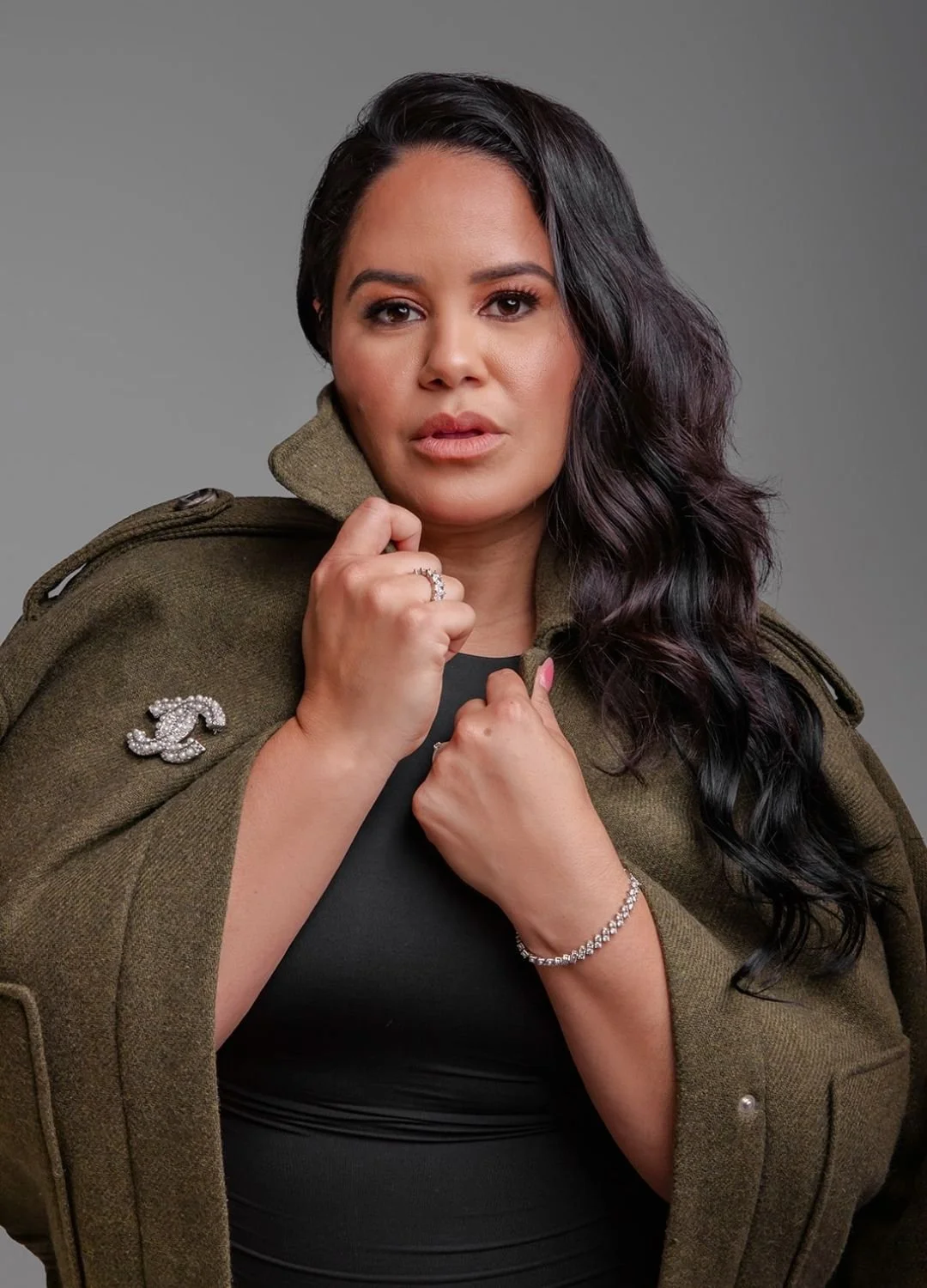 A woman with long dark wavy hair wearing a black top and an olive green jacket with a Chanel logo pin, standing against a gray background, holding her collar and looking directly at the camera.