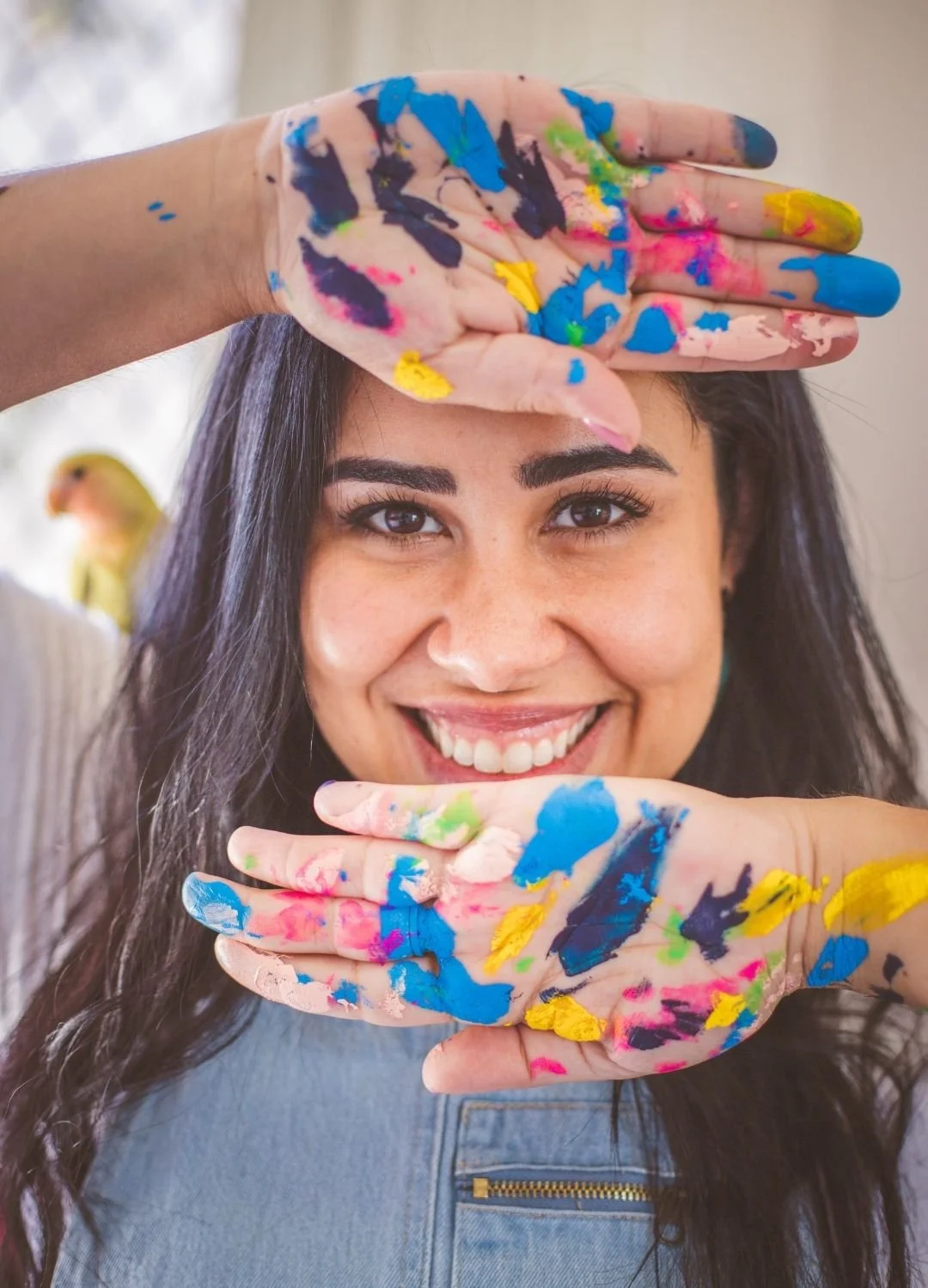 A smiling woman with dark hair and brown eyes, holding her painted hands in front of her face with bright colors of blue, pink, yellow, green, and white. She is wearing a gray shirt and is standing indoors with a blurred background.