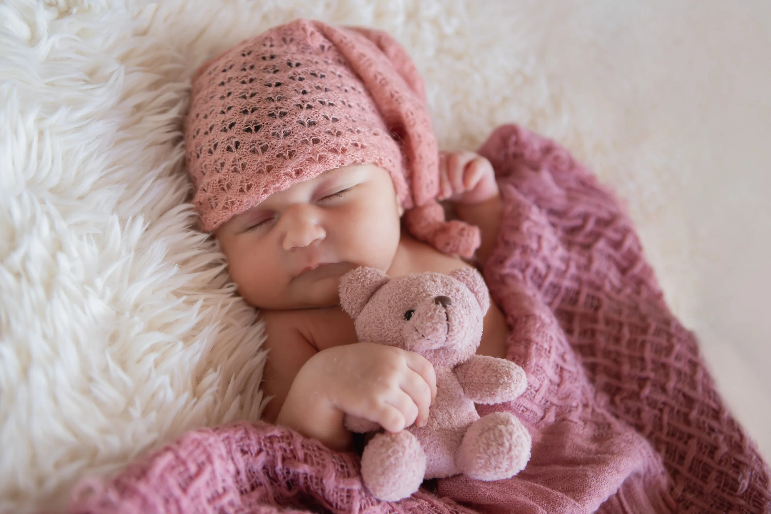 A sleeping baby with a pink knitted hat and matching blanket, holding a pink teddy bear on a fluffy white surface.