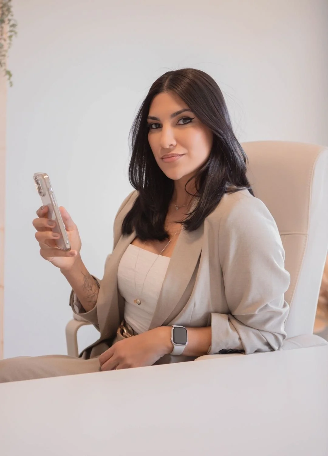 A woman with dark hair, wearing a beige blazer, holding a smartphone in her right hand, seated in a cream-colored chair, looking at the camera with a slight smile.