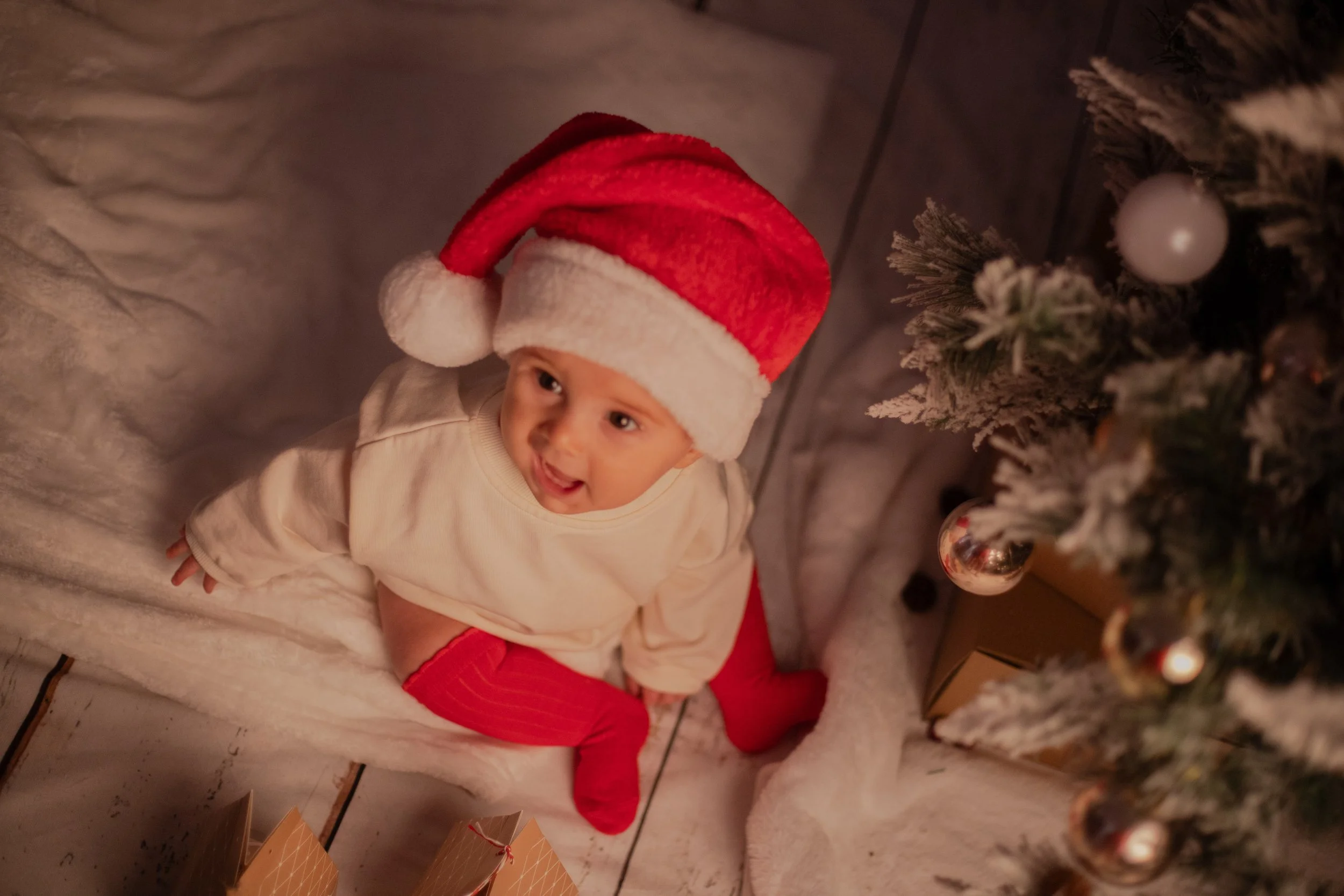 A baby wearing a Santa hat and red stockings, sitting on the floor near a decorated Christmas tree with wrapped gifts underneath.