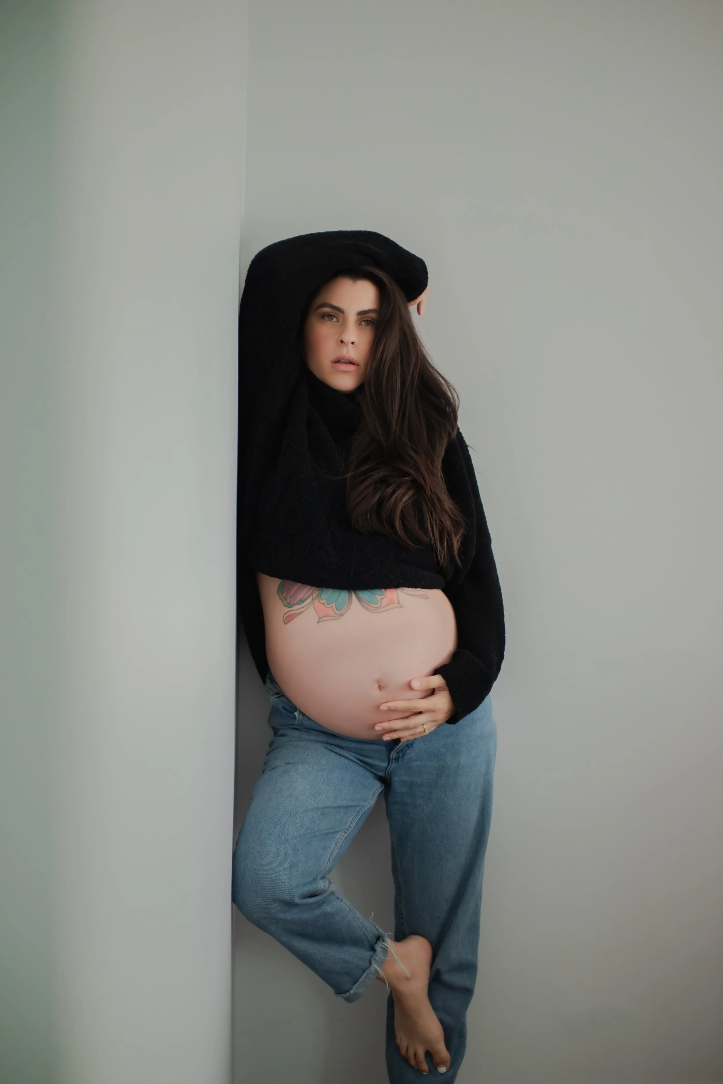 Pregnant woman with long dark hair, wearing a black hoodie and jeans, standing against a wall, with one hand on her belly and the other arm raised above her head, looking directly at the camera.