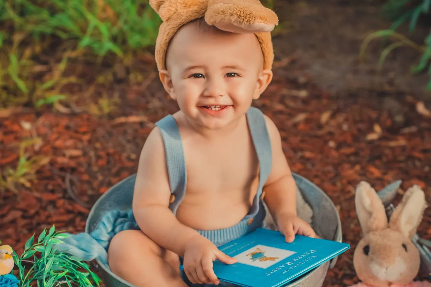 A smiling toddler with a bear hat, sitting in a basket outdoors, holding a book titled 'Fletcher and the Pirate' with a plush bunny beside him.