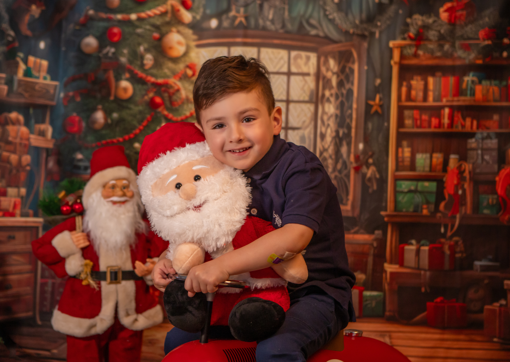 A young boy with brown hair riding a red toy tractor, hugging a plush Santa Claus, in a festive Christmas setting with Santa Claus, a decorated Christmas tree, and holiday decorations in the background.