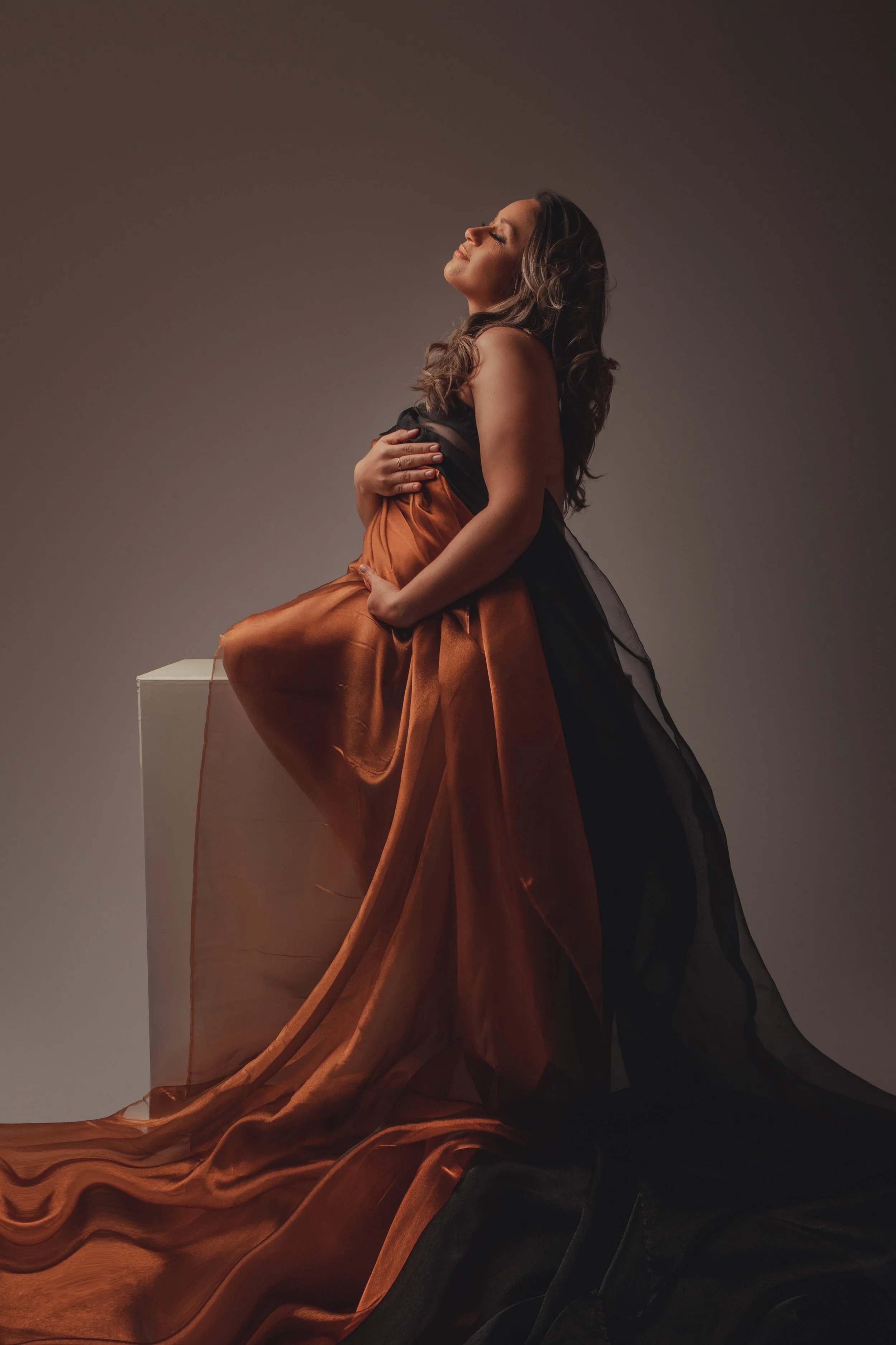 A woman in a flowing black and orange gown sitting with her head tilted back and eyes closed, holding her stomach. The gown has a layered, flowing design. She is seated on a white block against a neutral background.