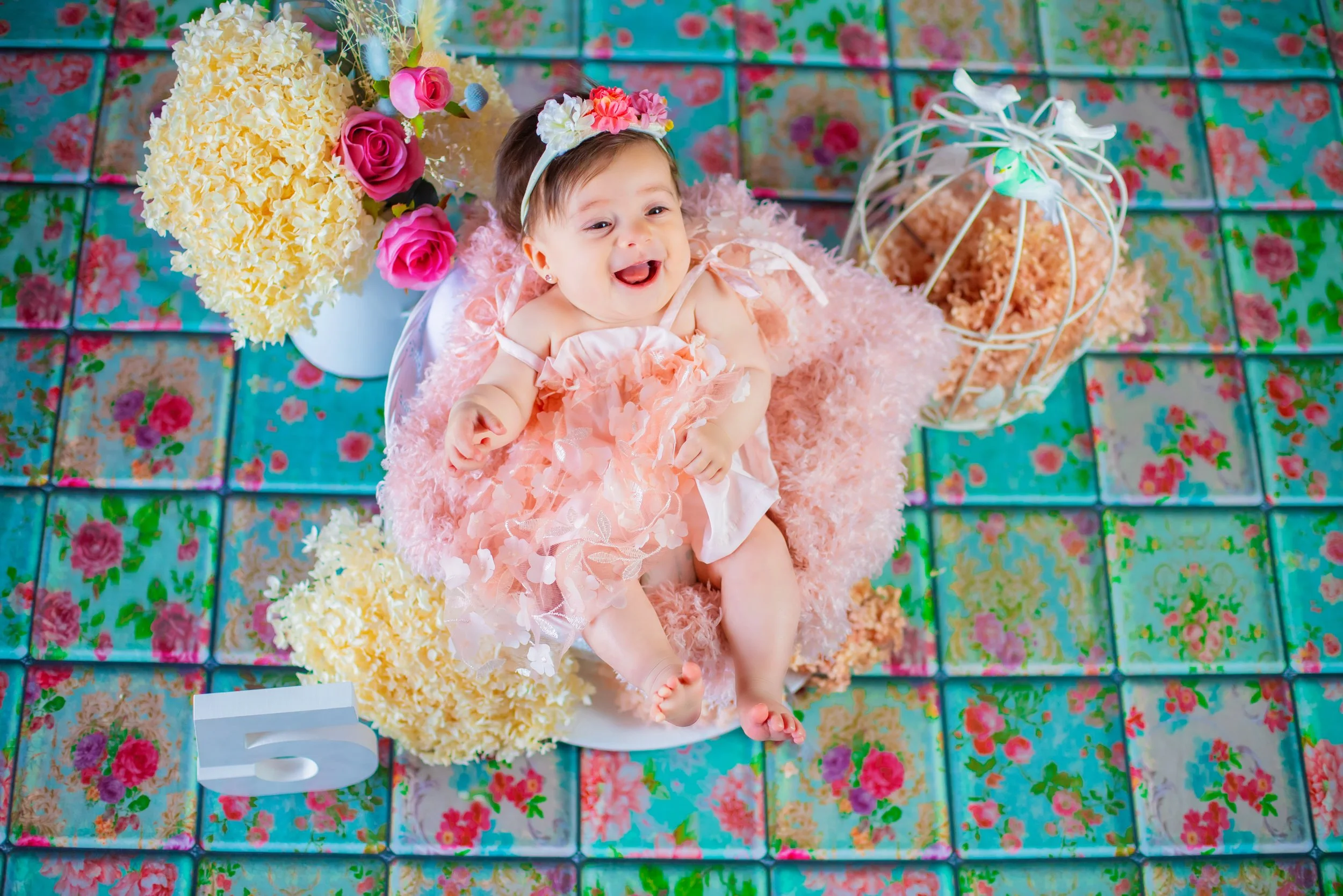 A smiling baby girl lying on a pink fluffy blanket surrounded by flowers, a white number five, and a white decorative birdcage with pink and white flowers inside, celebrating her fifth birthday.