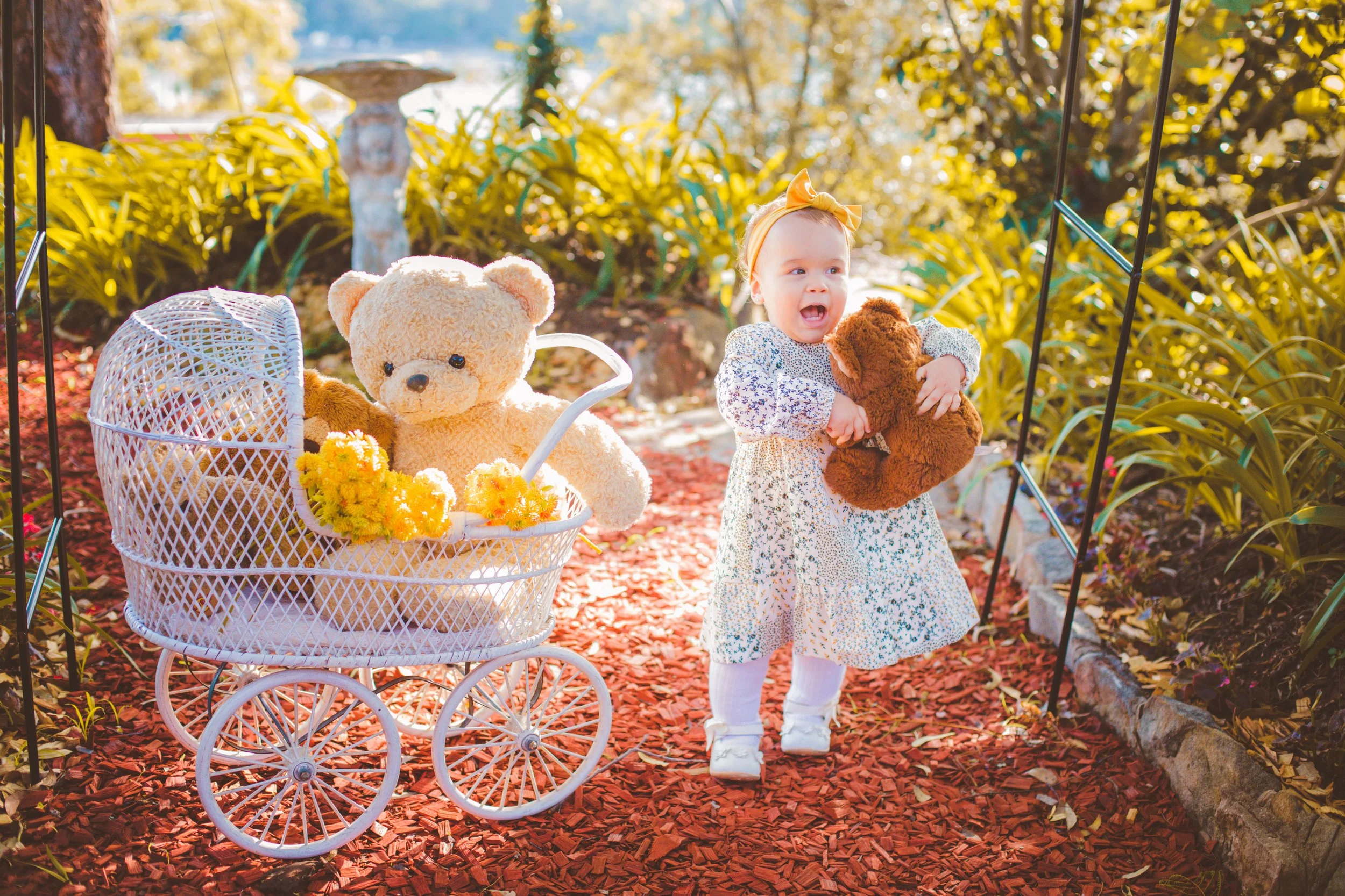 A young girl in a floral dress and yellow bow holding a teddy bear, standing on a red wood chip path, outdoors with lush green plants and flowers in the background, a white toy stroller filled with pink and yellow flowers and teddy bears to her left.