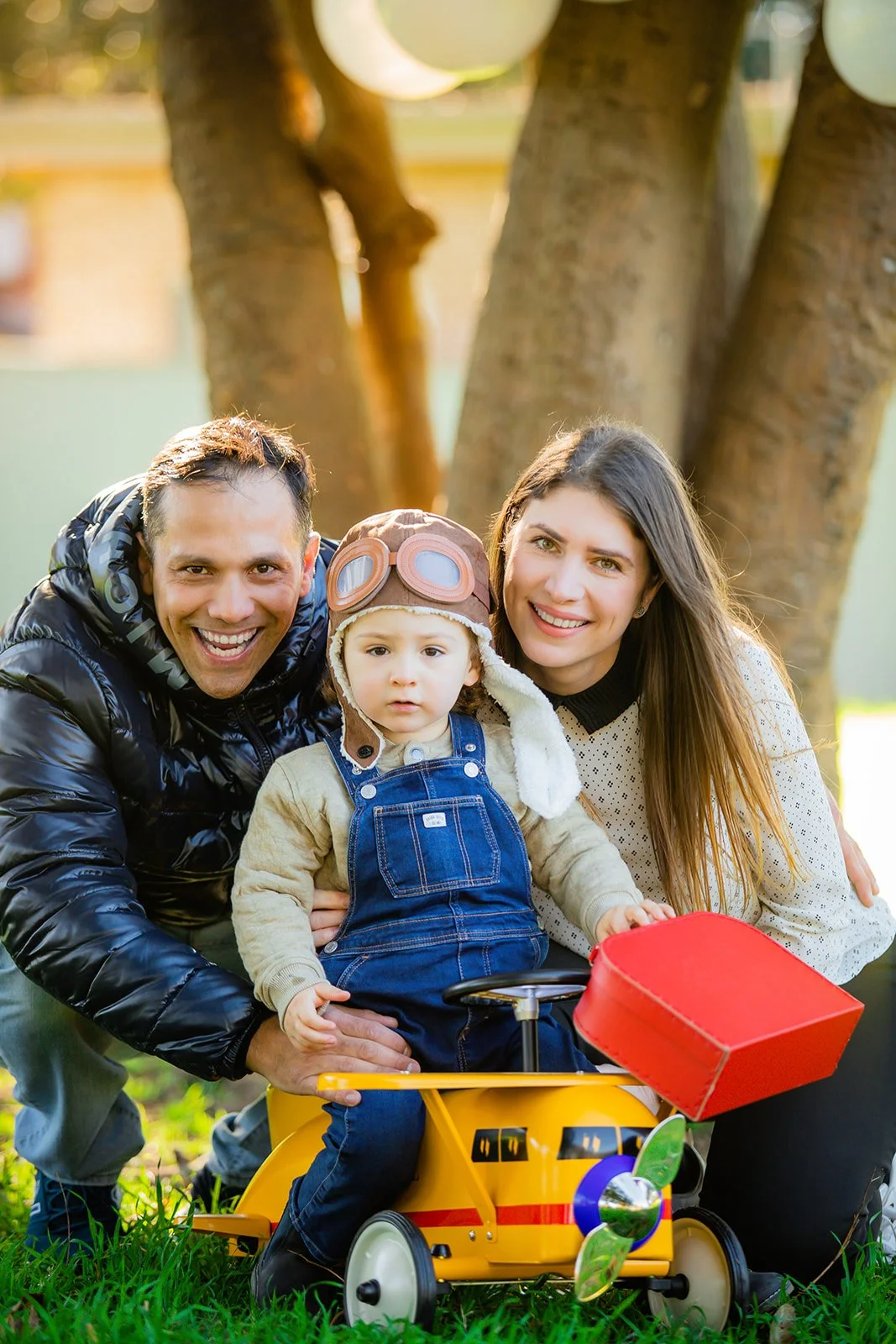 A happy family of three outdoors, with a man, woman, and young child dressed warmly, sitting on a grass field next to a tree. The child is sitting on a yellow toy race car, wearing goggles and a pilot's hat, with the parents smiling beside them.