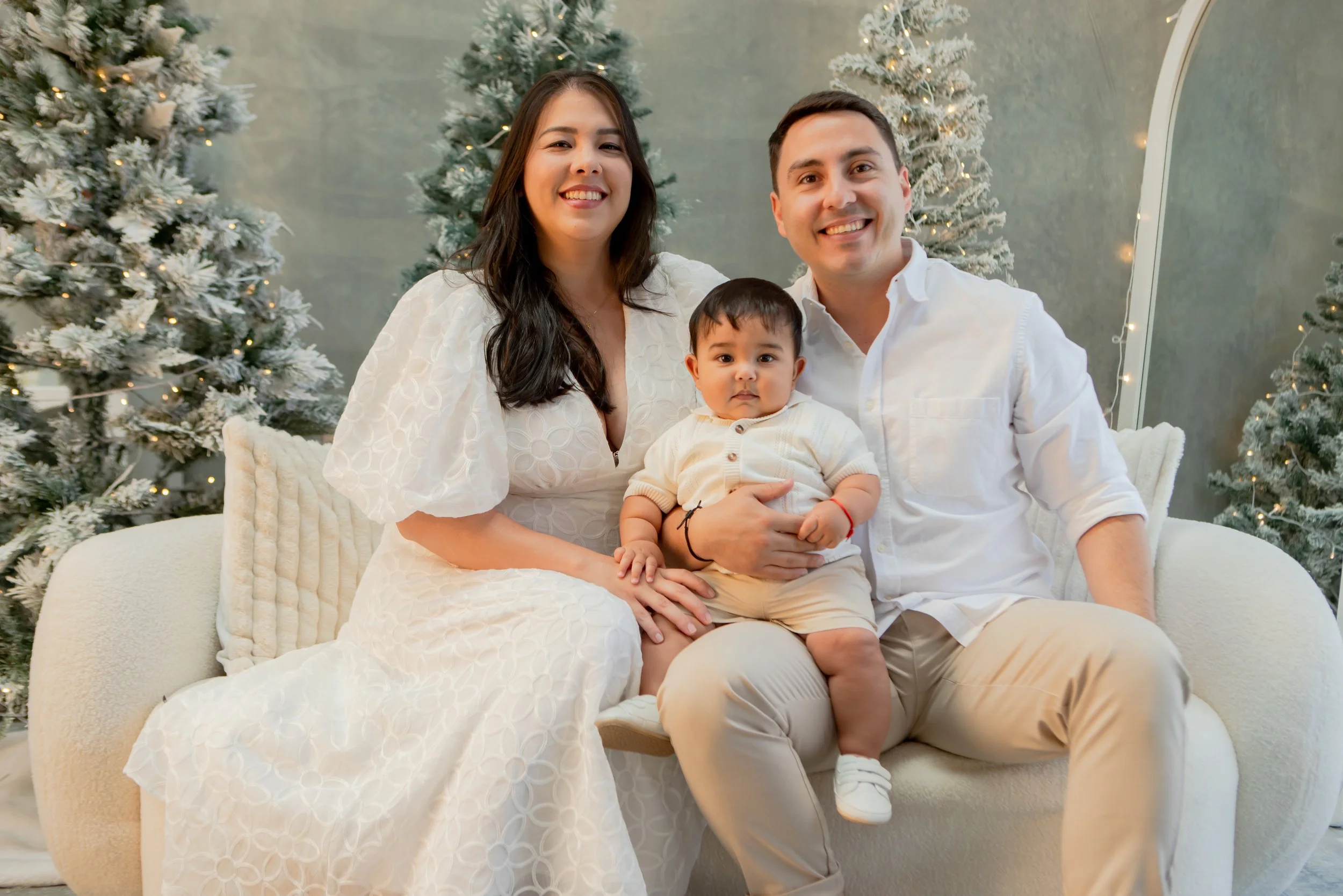 Happy family of three sitting on a cream-colored sofa, with Christmas trees decorated with lights in the background. The woman has long dark hair and is wearing a white dress, the man has short dark hair and is wearing a white shirt and beige pants, 