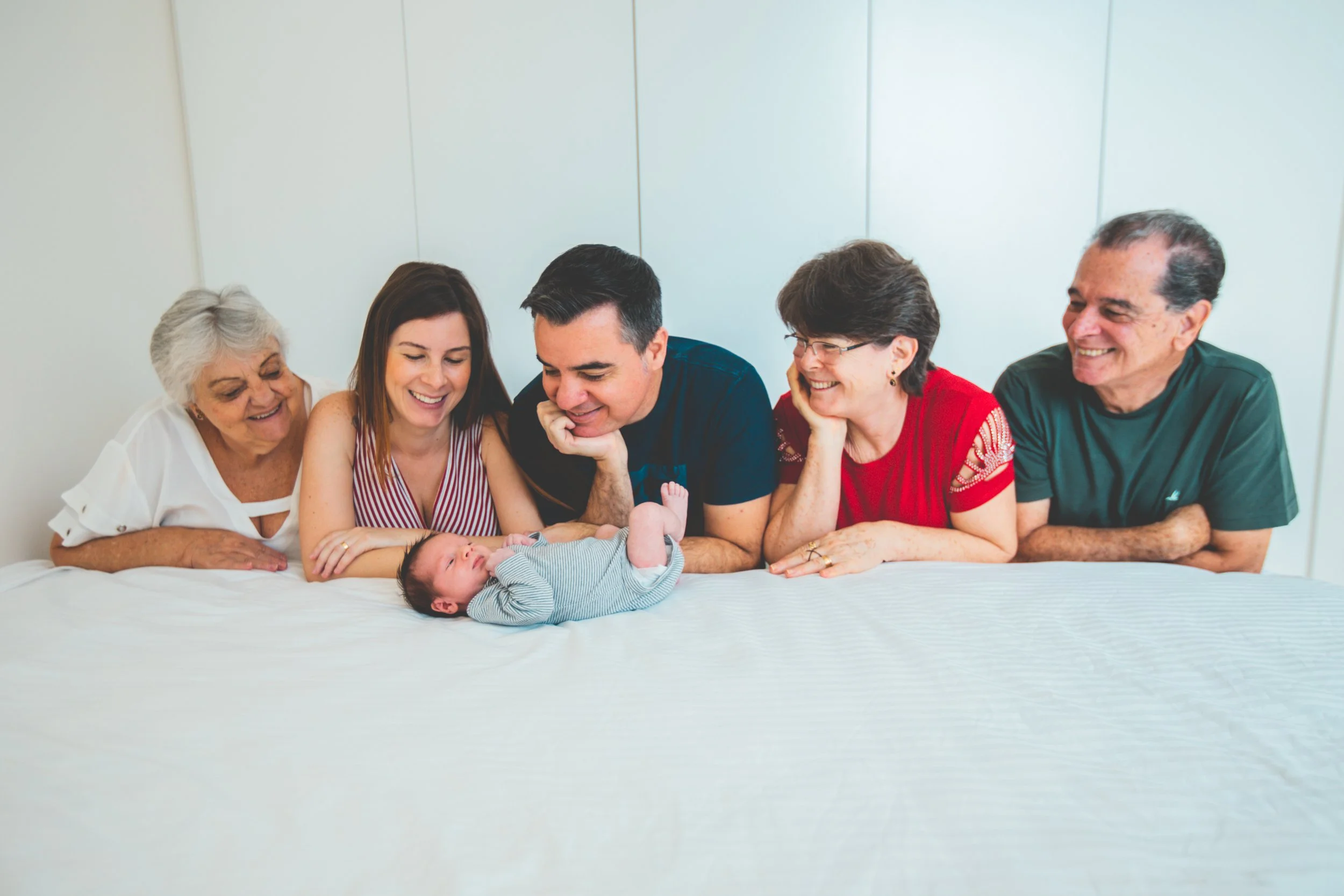 Family of six people, including three generations, gathered around a newborn on a bed, smiling and looking at the baby.