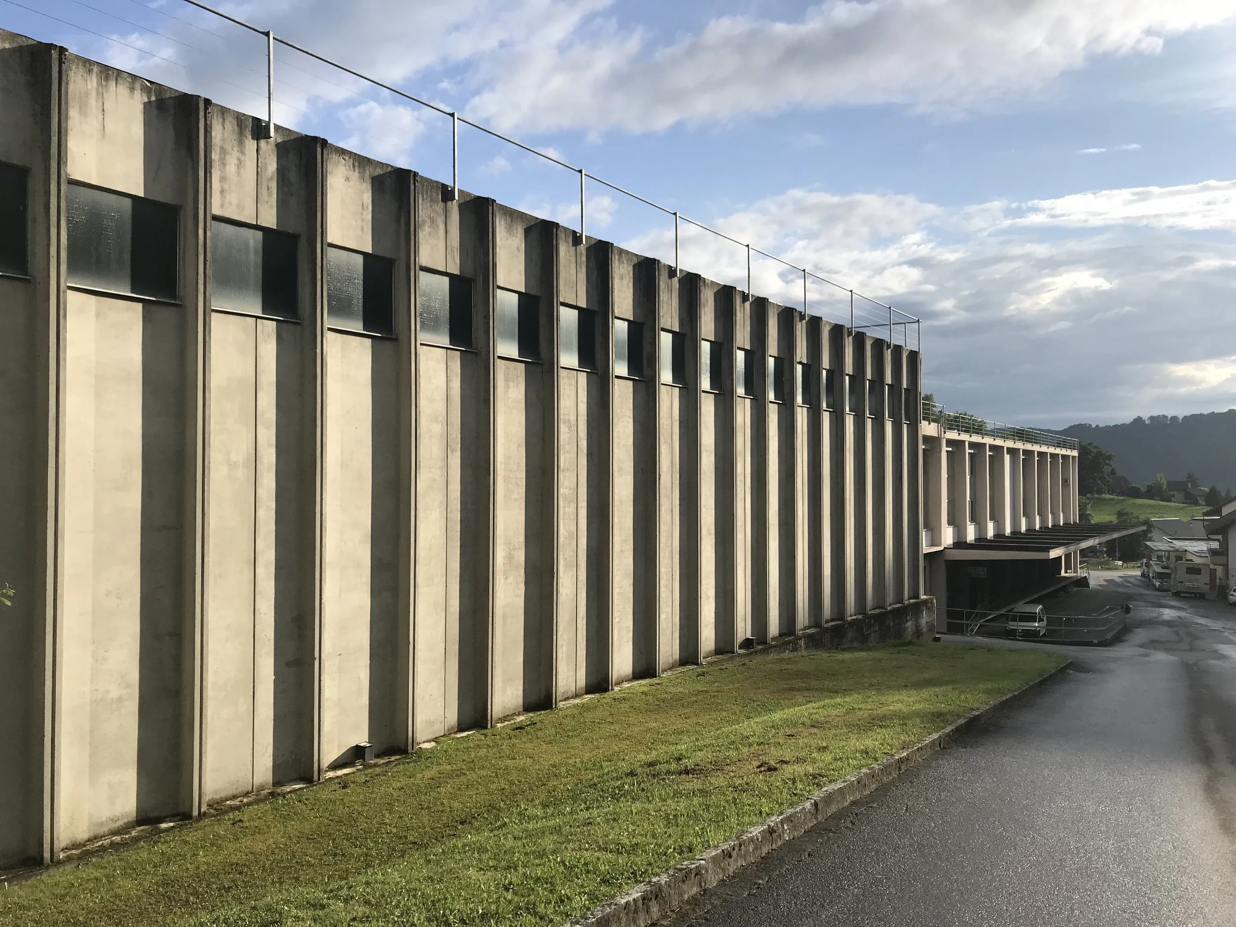 Bâtiment moderne en béton avec plusieurs fenêtres, ciel nuageux, route mouillée et espaces verts.