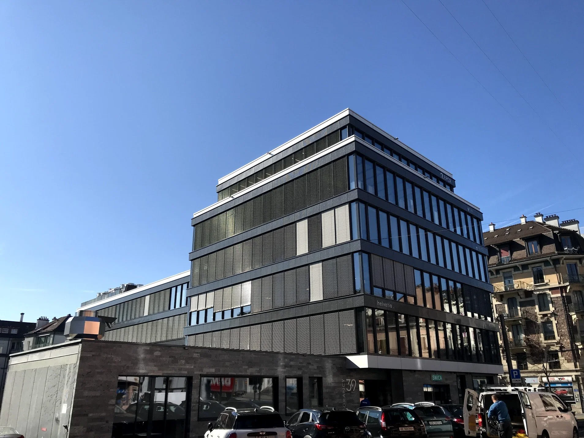 Immeuble moderne à plusieurs étages avec façade en verre et des panneaux métalliques, situé dans une zone urbaine avec des voitures garées devant et des bâtiments résidentiels à l'arrière.