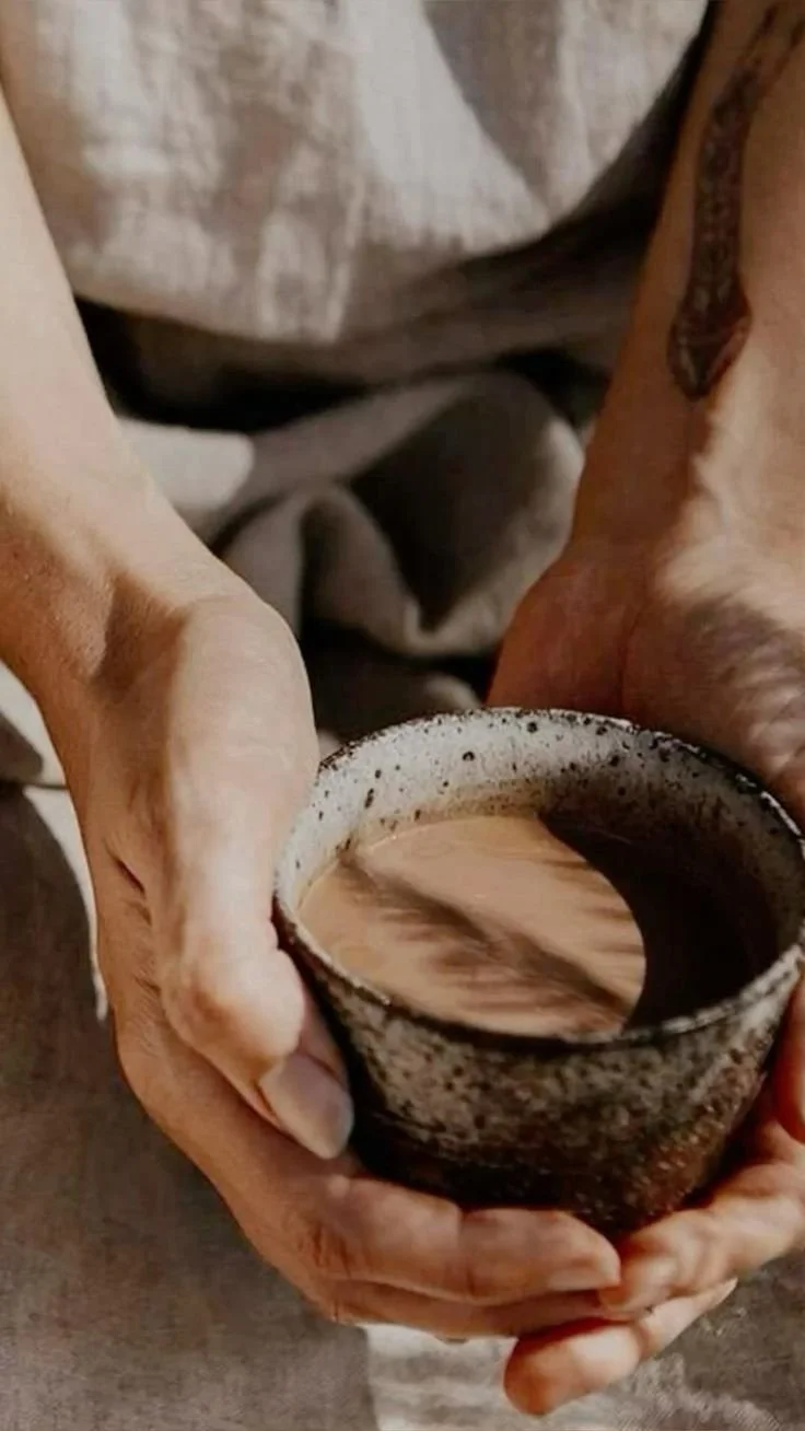 A person holding a ceramic mug filled with a frothy, brown beverage, possibly coffee or hot chocolate, with a swirling pattern on the surface.