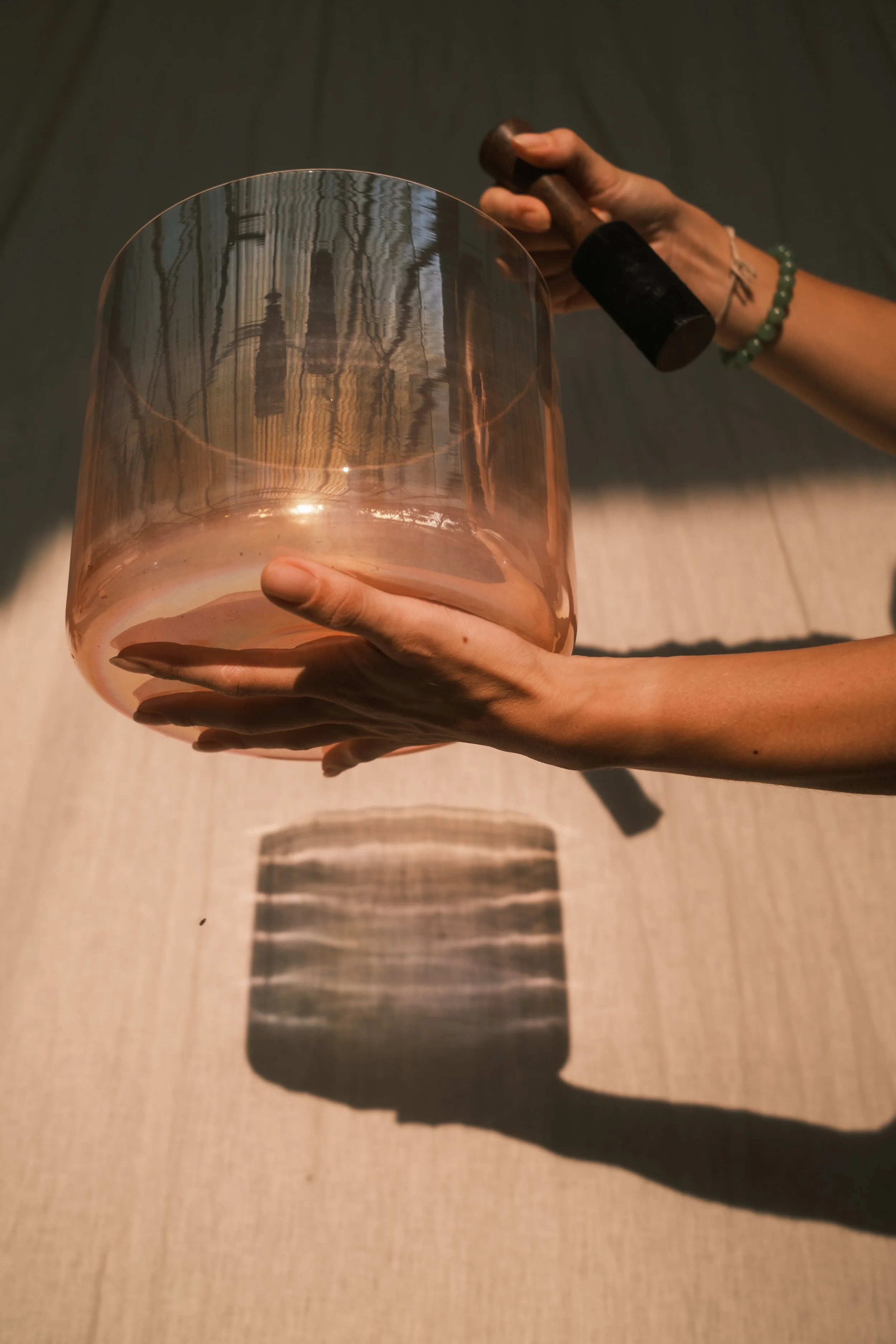 Person holding a large, transparent, pinkish glass vase with their hands, casting a striped shadow on the floor.