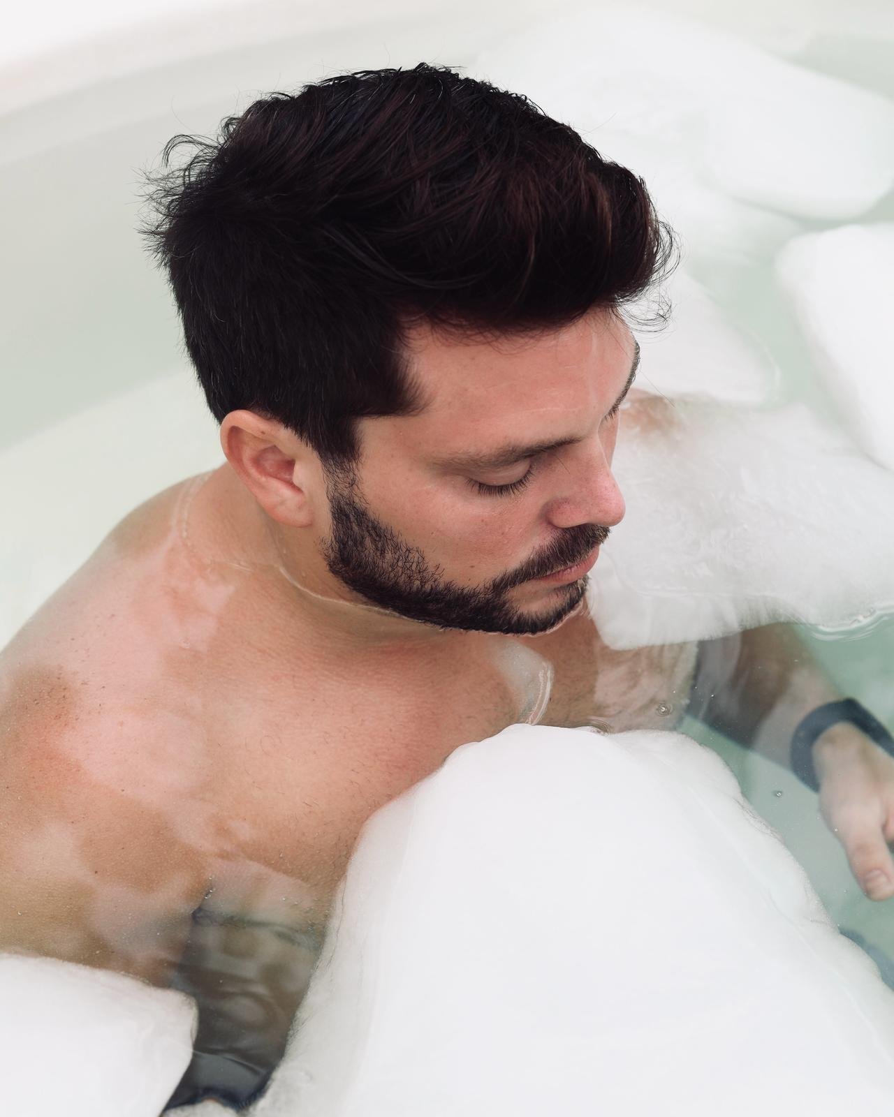 A shirtless man with dark hair and a beard relaxes in a hot tub, with his eyes closed, surrounded by white foam.