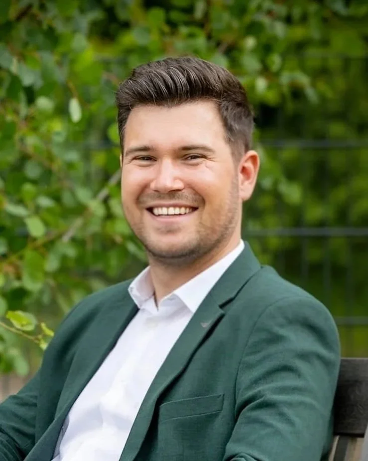 Ein lächelnder Mann in einem weißen Hemd und grüner Jacke sitzt auf einer Parkbank vor grünem Laub. Der Mann heißt Lucas Best.