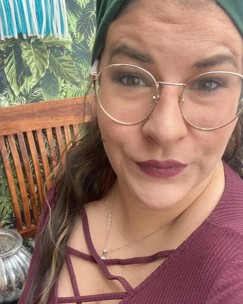 Nahaufnahme einer Frau mit Brille, dunklem Lippenstift, grüner Kopfbedeckung, im Hintergrund grüne Pflanzen und ein Holzstuhl. Die Frau heißt Tamara Müller.