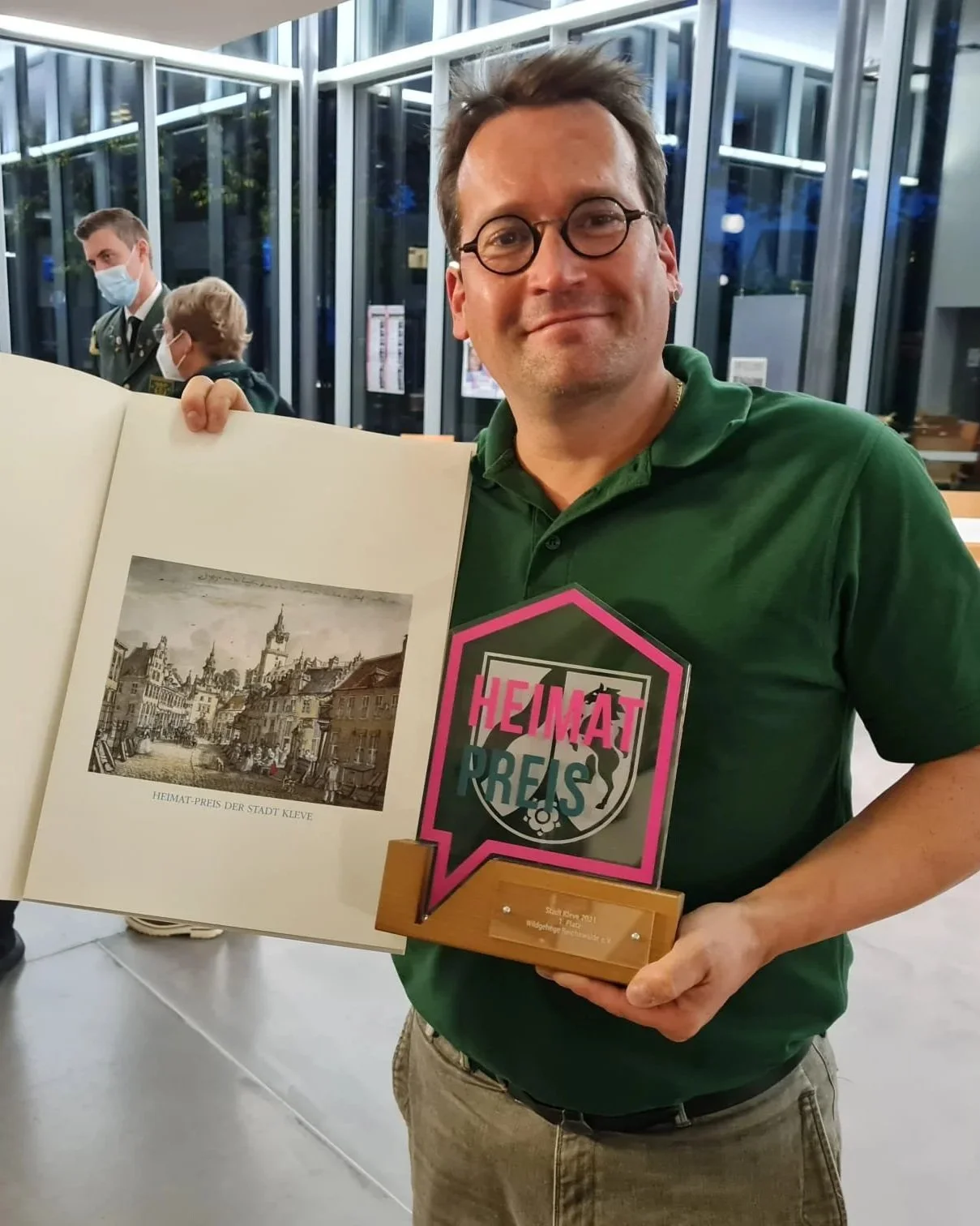 Ein lachender Mann in einer grünen Polo trägt eine Trophäe mit der Aufschrift »Heimatpreis«. Er hält ein offenes Buch, in dem eine alte schwarz-weiße Stadtaufnahme mit der Beschriftung »Heimat-Preis der Stadt Kleve« zu sehen ist. Im Hintergrund sind zwei Männer mit Gesichtsmasken im Gespräch.