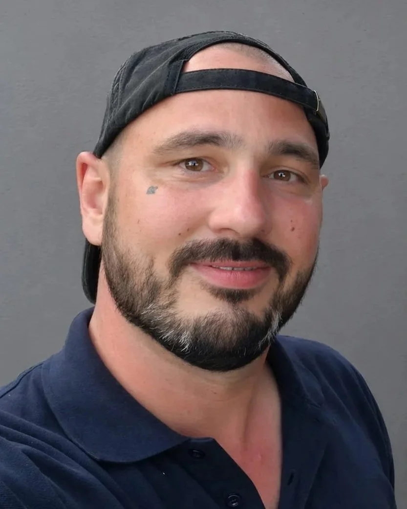 Nahaufnahme eines lachenden Mannes mit Bart, Barthaare, dunklem T-Shirt und schwarzer Baseballcap vor grauem Hintergrund. Der Mann heißt Maik Wolters.