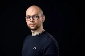 Portrait of a man with glasses, a beard, and a mustache, wearing a navy blue shirt, against a black background.