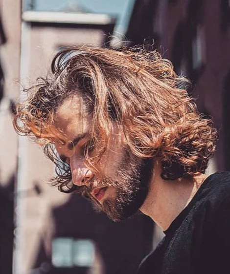 Profile of a man with curly, reddish-brown hair and a beard, outdoors in sunlight.