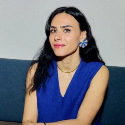 Woman with long dark hair wearing a blue sleeveless top and large blue earrings, sitting on a dark-colored couch against a plain background.