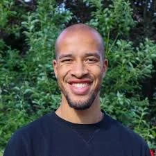 Portrait of a man with glasses, a beard, and a mustache, wearing a navy blue shirt, against a black background.