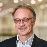 A middle-aged man with glasses, smiling, wearing a dark blazer and a light-colored shirt, standing in an indoor setting with blurred bright lights in the background.