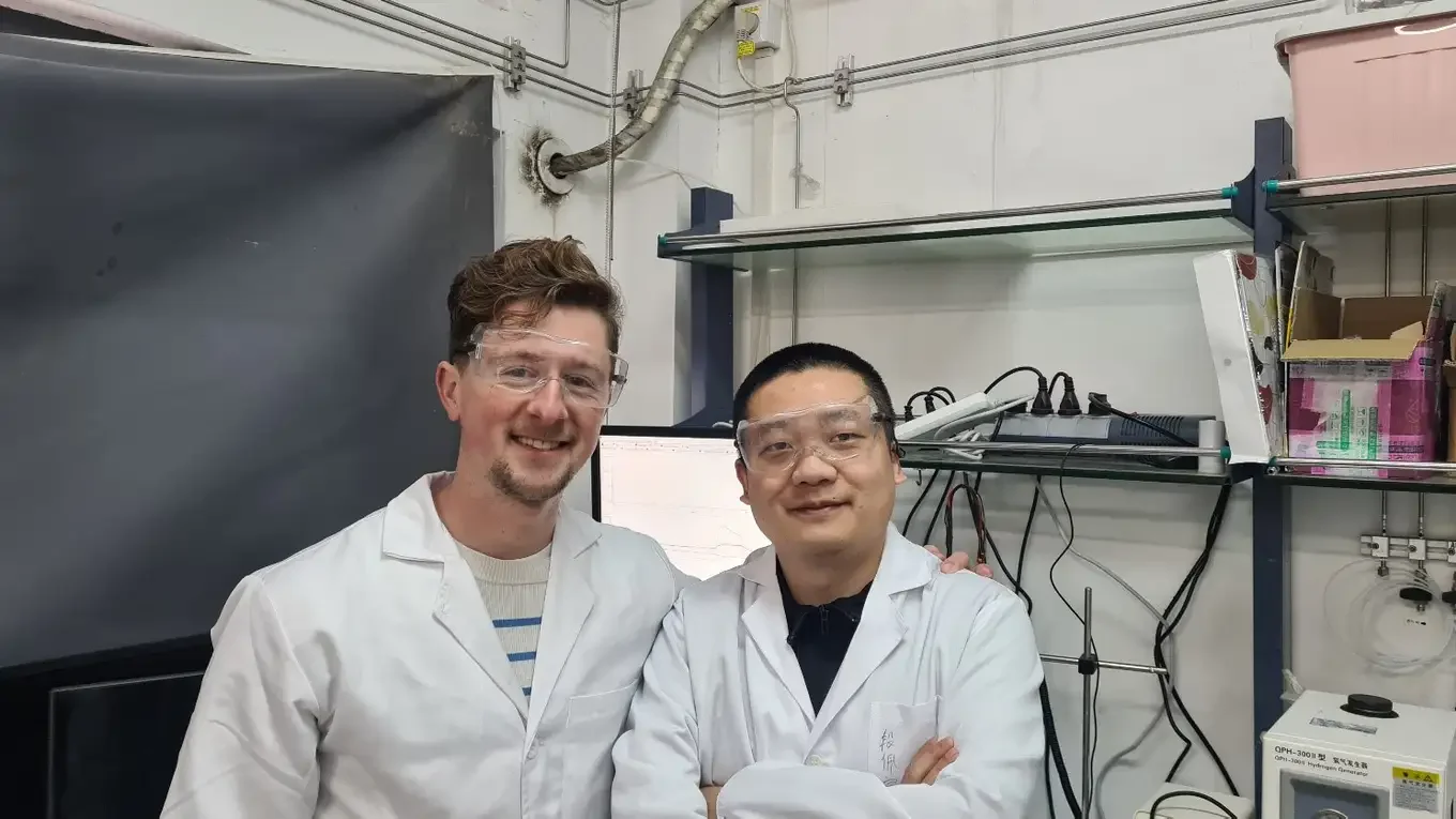 Two scientists in lab coats and safety goggles standing together in a laboratory.
