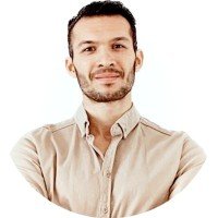 A young man with short dark hair and a beard, smiling with arms crossed, wearing a beige button-up shirt against a white background.