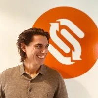 Person with curly hair smiling, standing in front of an orange and white logo or sign.