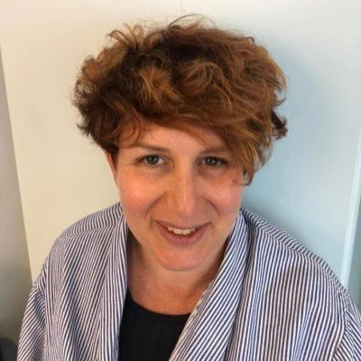 Portrait of a middle-aged woman with short blond hair, wearing a navy blazer and earrings, standing and smiling in an indoor setting.
