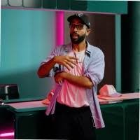 A man with a beard and glasses wearing a hat, grey shirt, and white t-shirt underneath, standing in a room with neon pink and teal lighting, with a phone and other objects on a counter behind him.