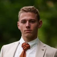 Young man in a beige suit with a brown tie, outdoors against a green background.